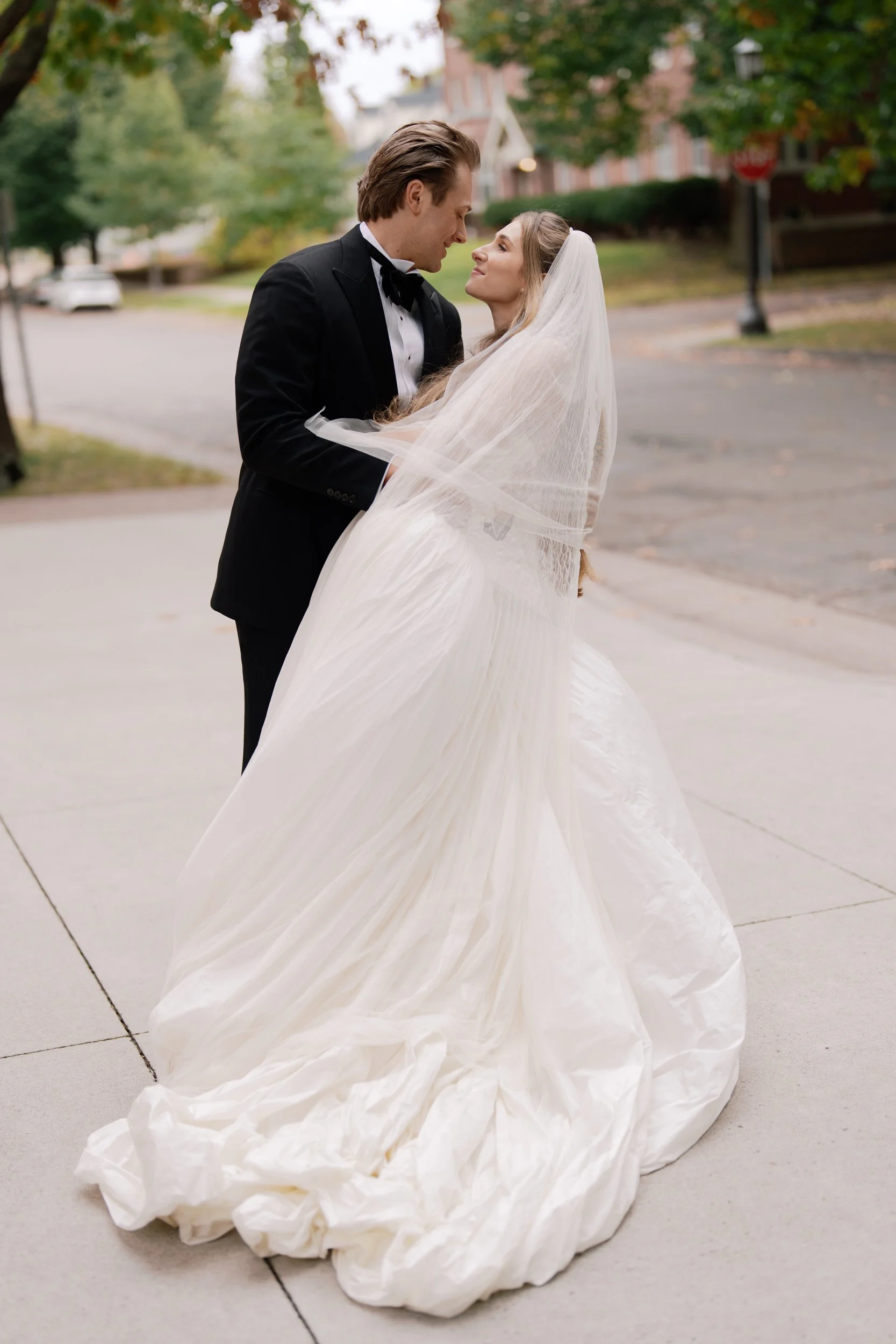 A man in a black tuxedo and a woman in a white wedding gown and veil standing outdoors, gazing into each other's eyes on the sidewalk with trees and houses in the background.