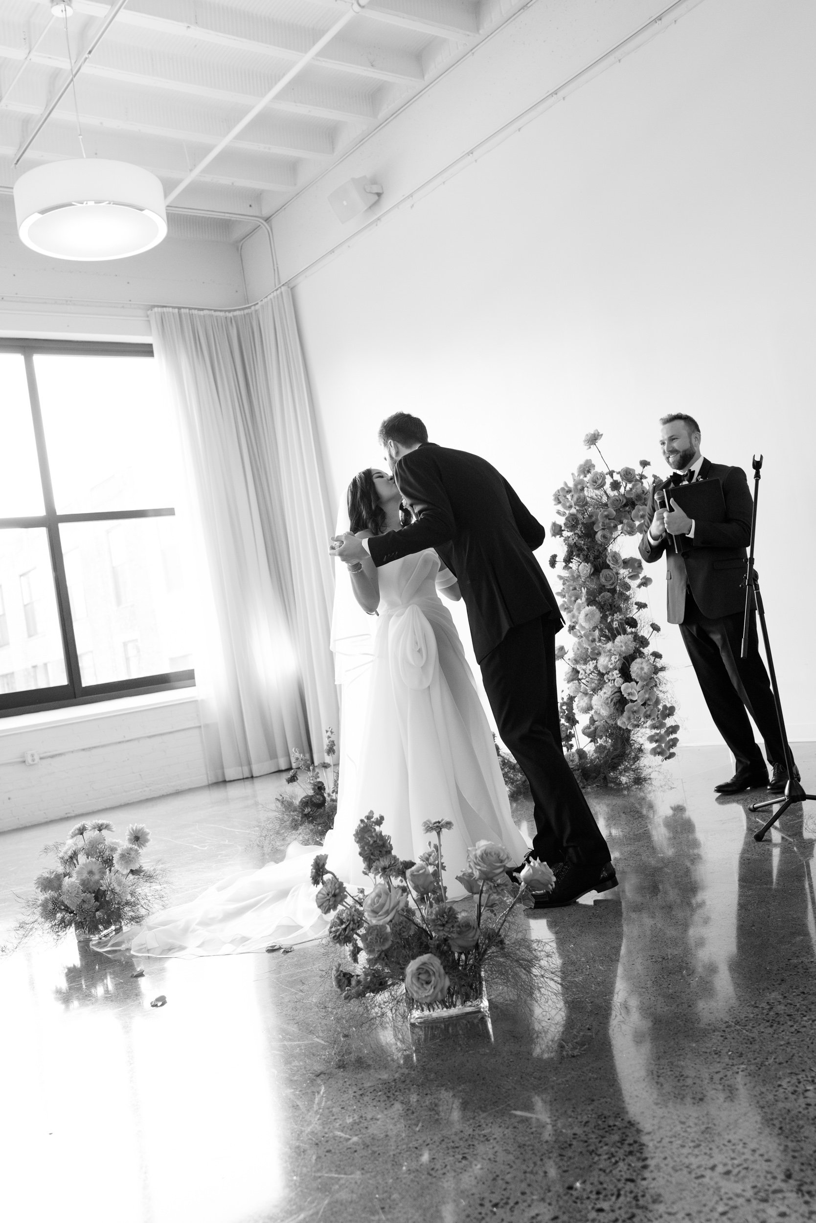Black and white photo of a wedding ceremony with a bride and groom kissing, an officiant holding a book, and floral arrangements, in a bright room with large window and curtains.