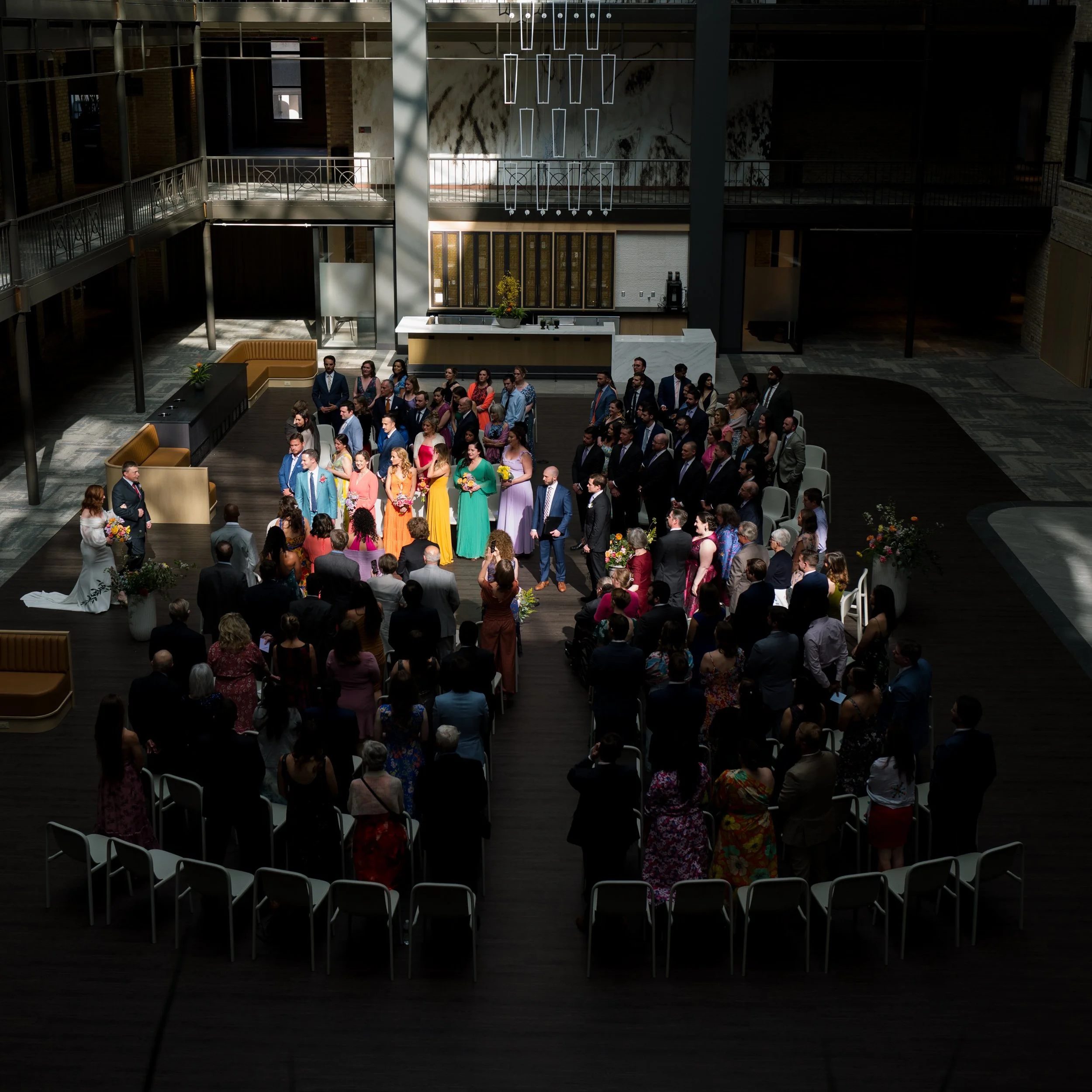 A wedding ceremony taking place in a modern indoor venue with a large group of guests gathered around, facing a bride and groom at the front. The venue features high ceilings, contemporary decor, and natural light.