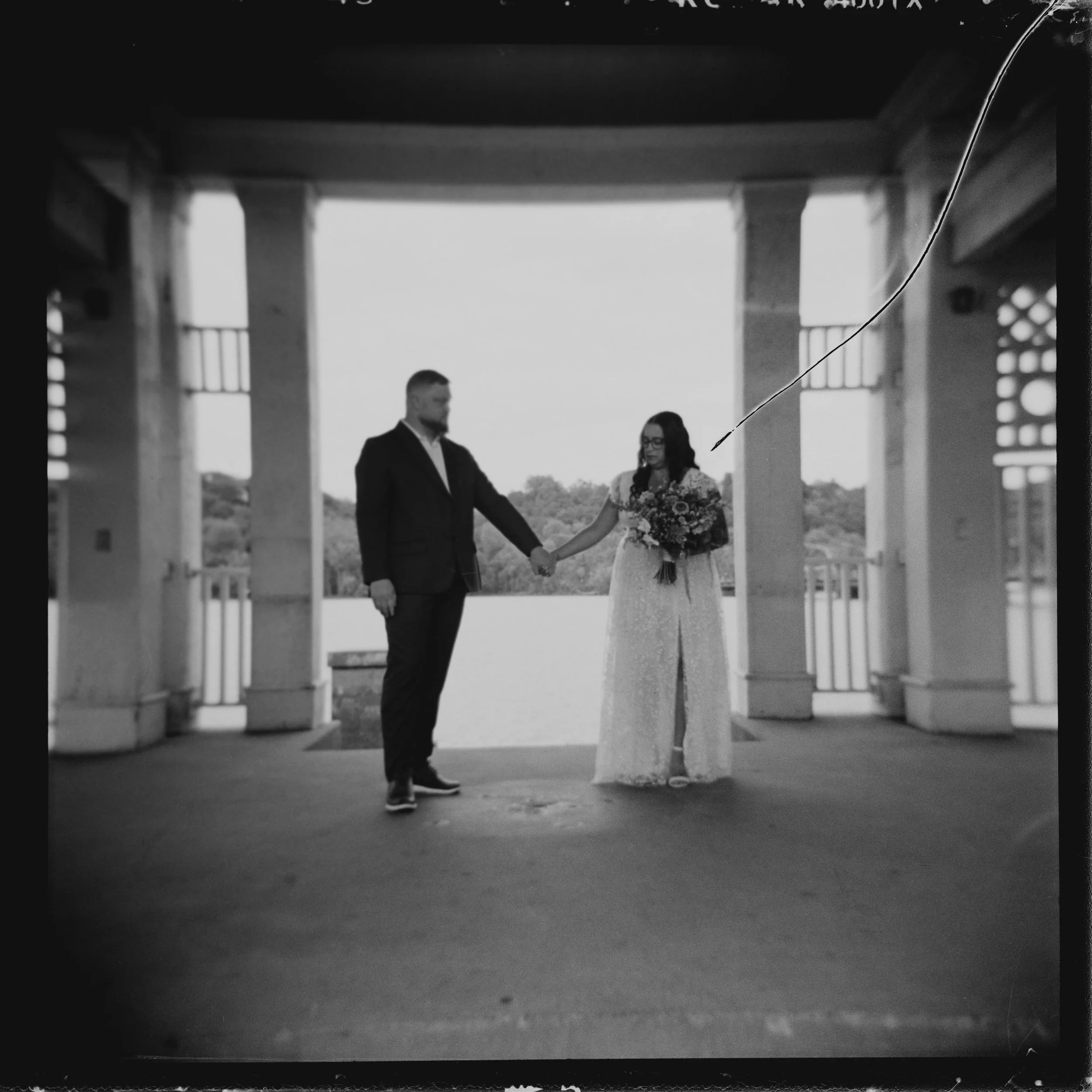 A black-and-white photo of a couple holding hands under a pavilion, with water and trees in the background. The woman is holding a bouquet and wears a long dress, while the man is in a suit.