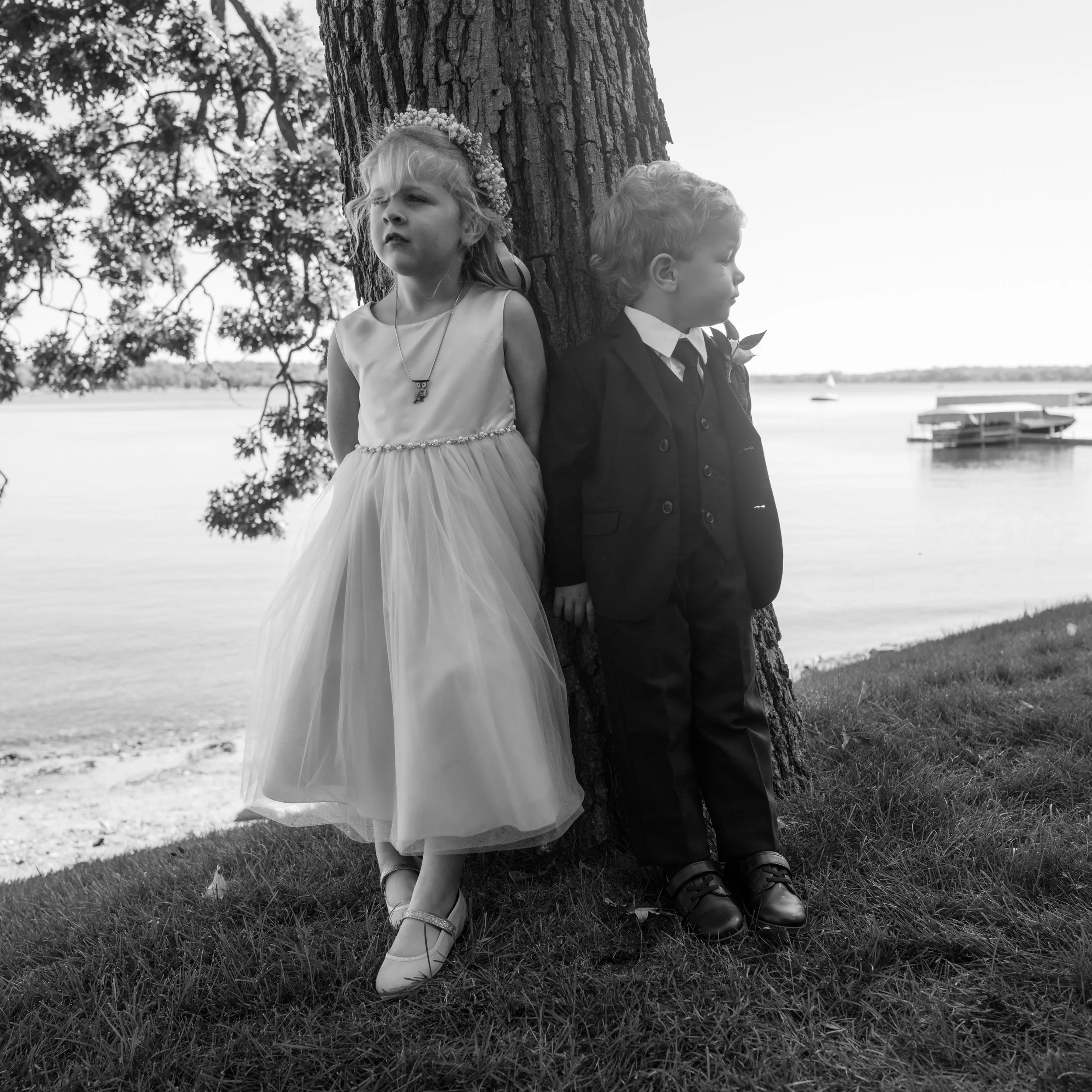 A young girl in a light-colored dress and a young boy in a suit stand back to back against a tree near a body of water, looking away from each other, during daytime.
