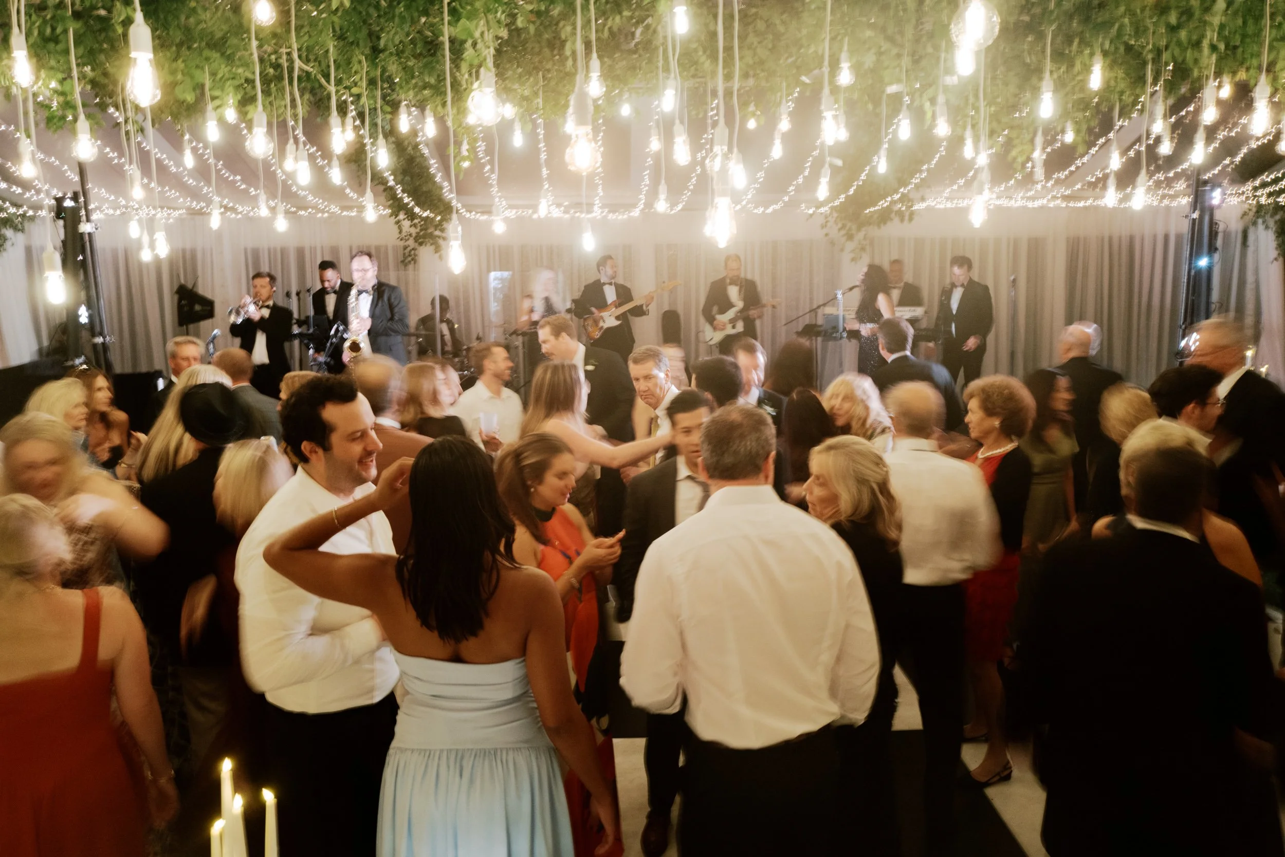 Crowd of people dancing at a formal event with a live band playing on stage, decorated with hanging lights.