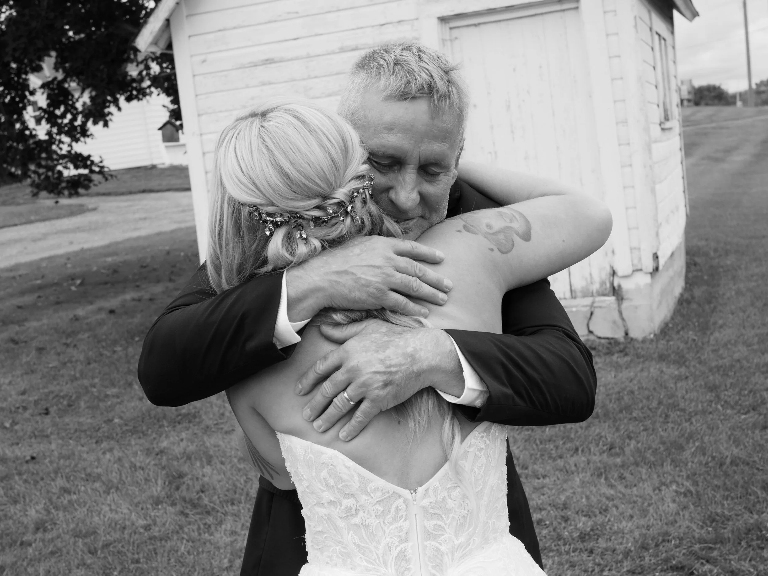 A black and white photo showing a man and a woman hugging closely outdoors. The woman is wearing a wedding dress and has a floral hair accessory, suggesting a wedding. The man is wearing a suit and there is a white wooden building in the background.