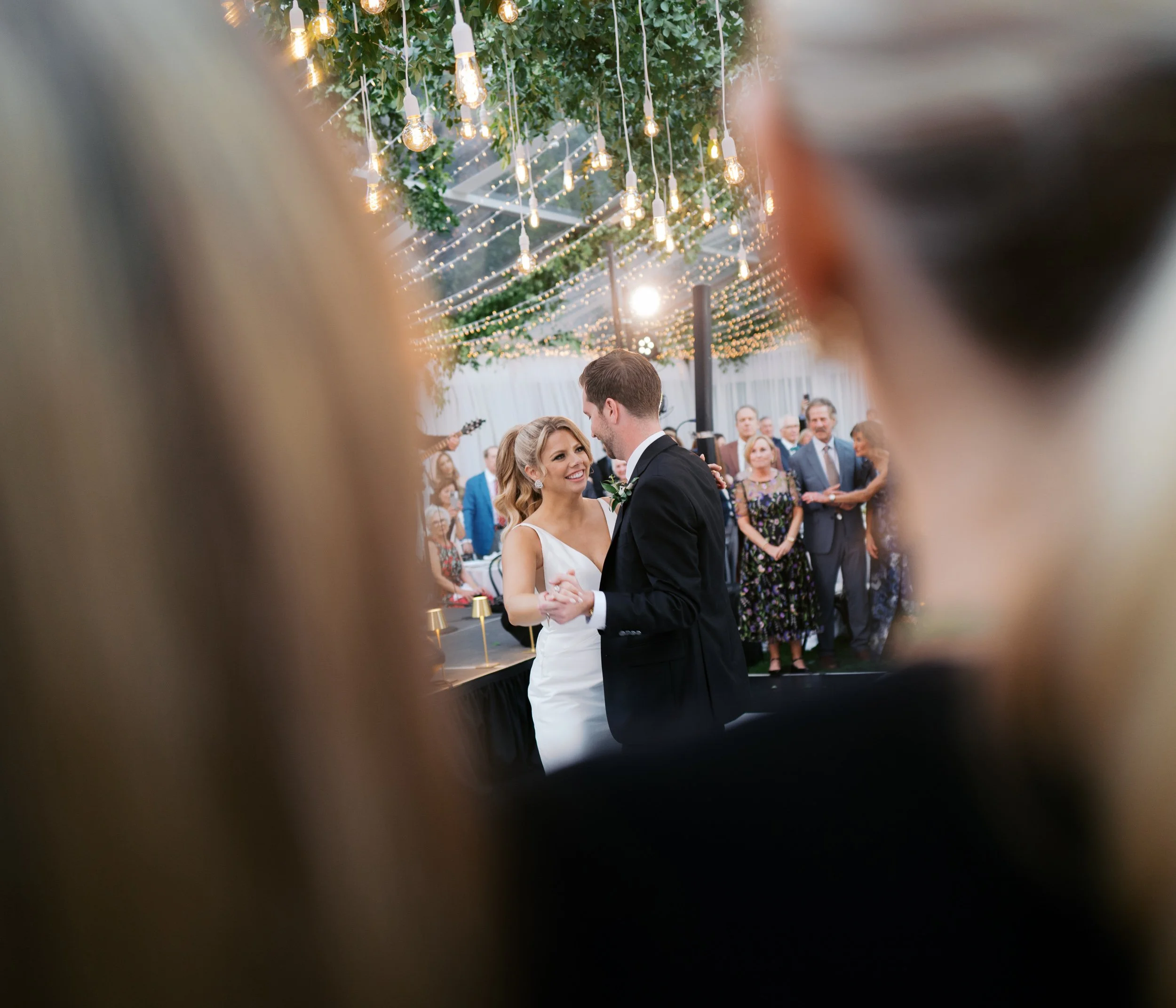 A bride and groom sharing their first dance at a wedding reception, framed by out-of-focus guests in the foreground, with string lights and hanging bulbs decorating the ceiling.