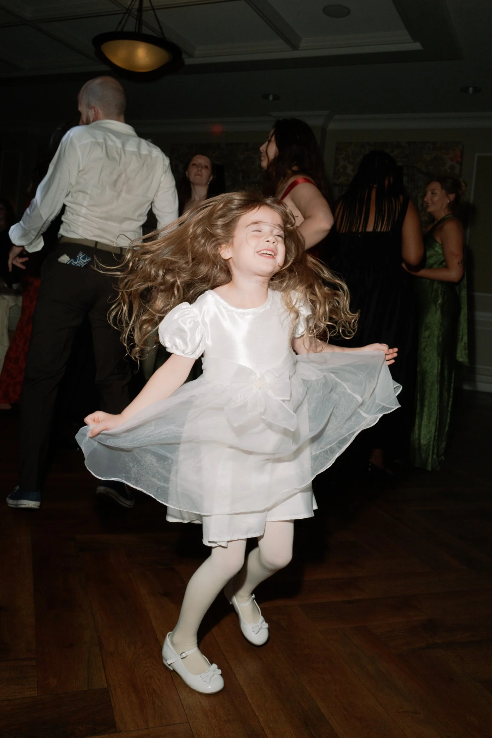 A young girl with long, wavy blonde hair dancing in a white dress with puffed sleeves and a bow, smiling with her eyes closed, on a dark wooden floor at a party or celebration with other adults in the background.