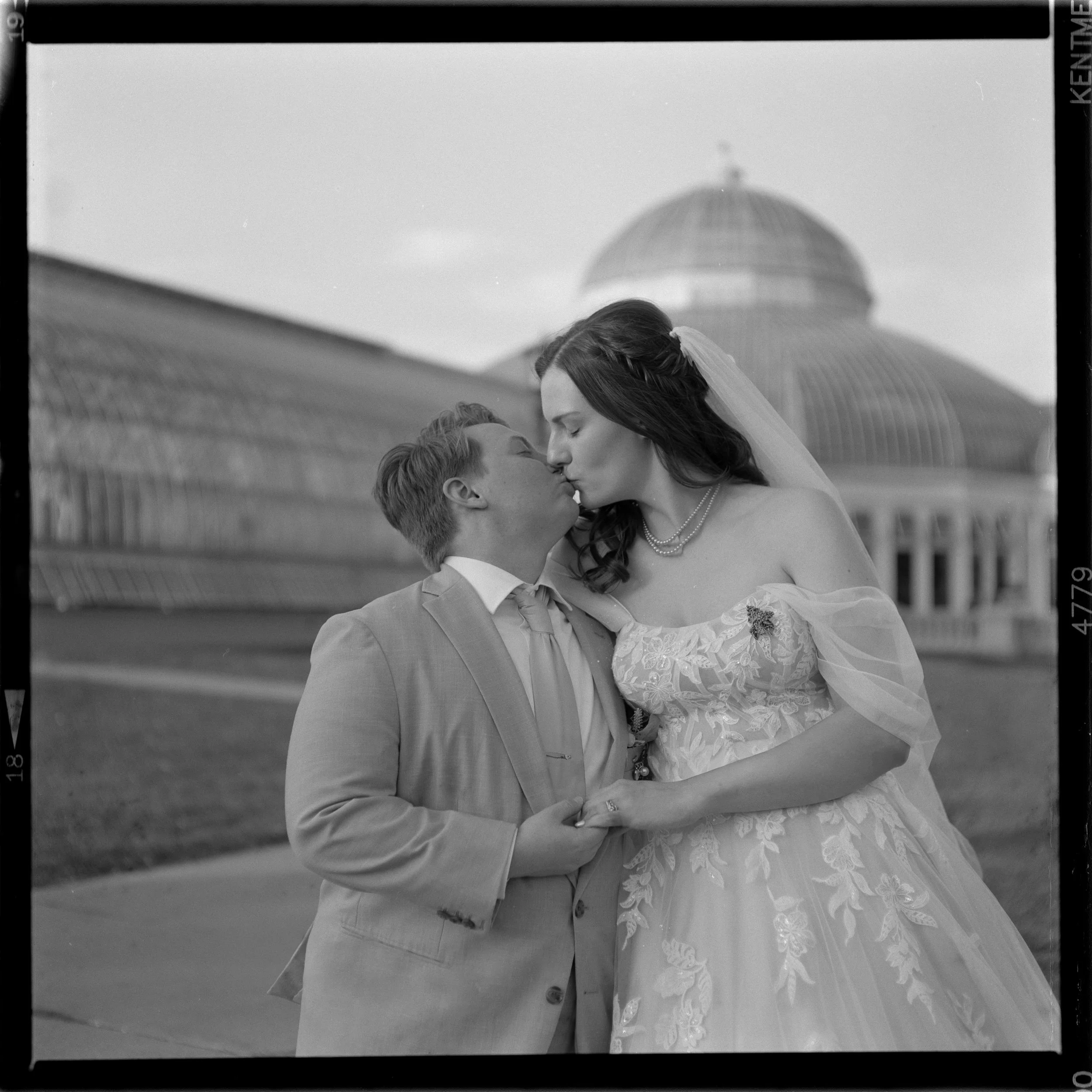 A black and white photo of a bride wearing a wedding gown with floral embroidery and a veil, and a groom in a suit, sharing a kiss outdoors with a domed building in the background.