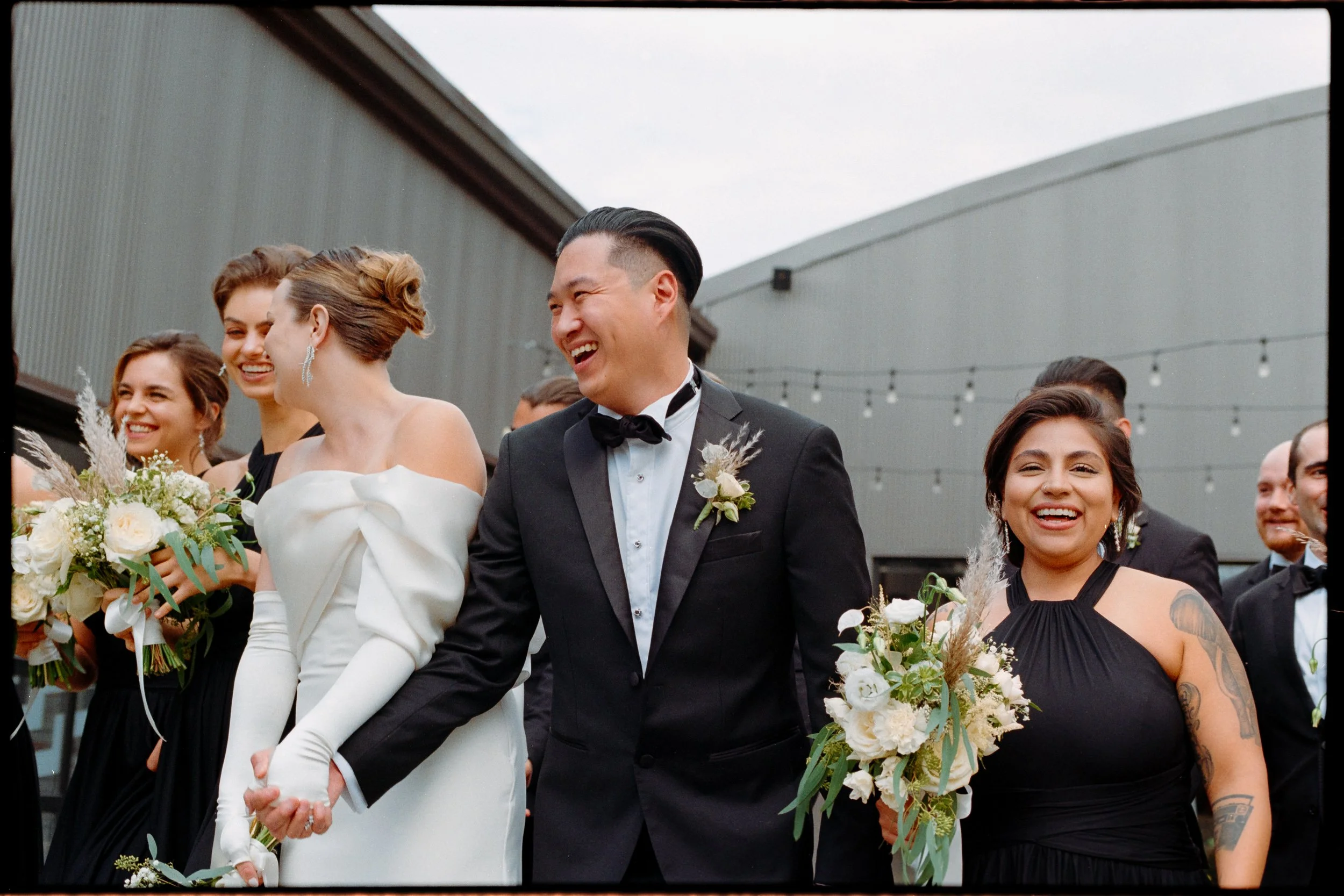 A joyful wedding party walking outdoors, with a diverse group of women in elegant dresses and a man in a tuxedo, holding bouquets of white flowers and laughing together.