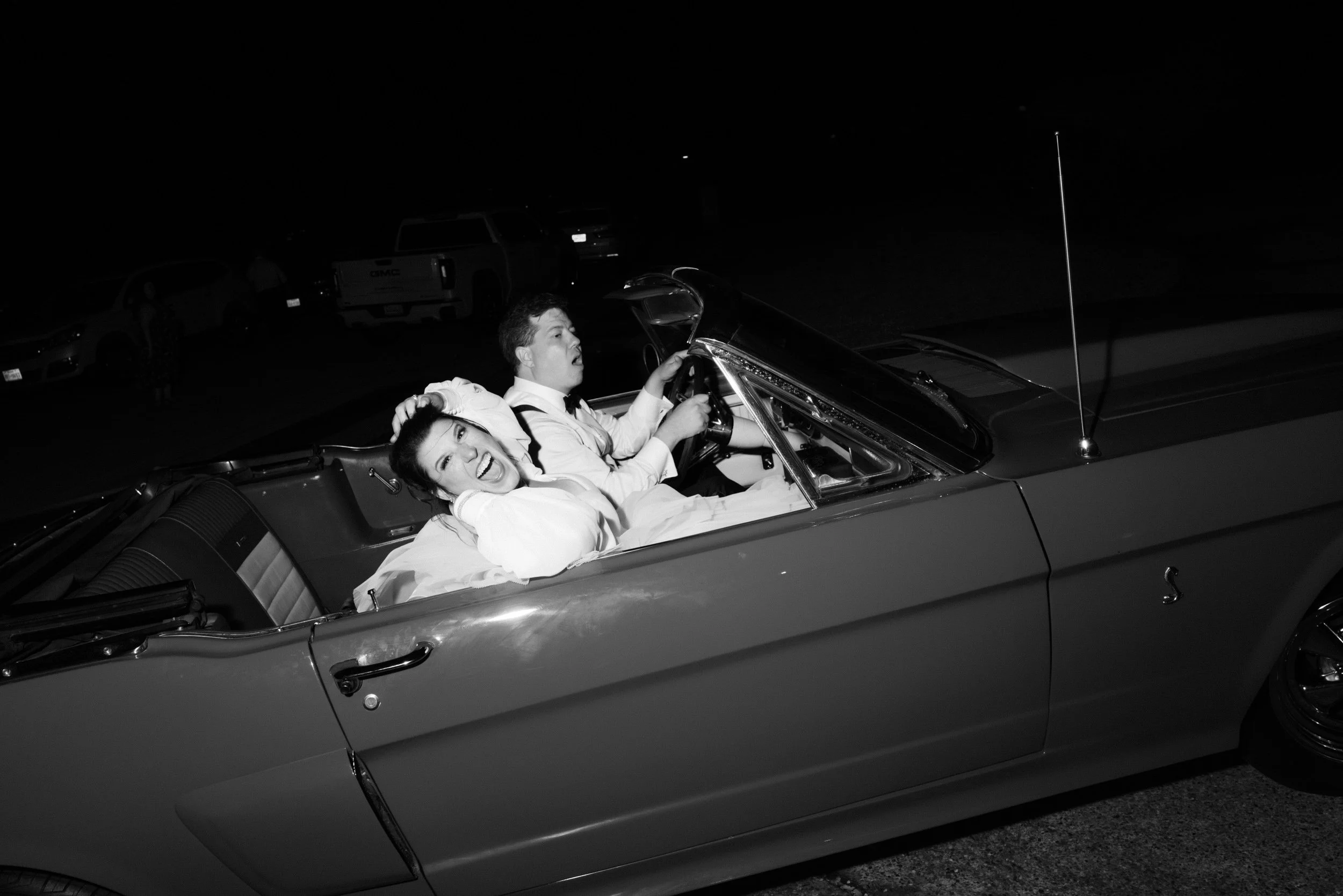 Black and white photo of a woman and a man in a convertible car at night; the woman is in the passenger seat, smiling and posing with hands near her head, while the man is driving, wearing a shirt and suspenders, looking ahead.
