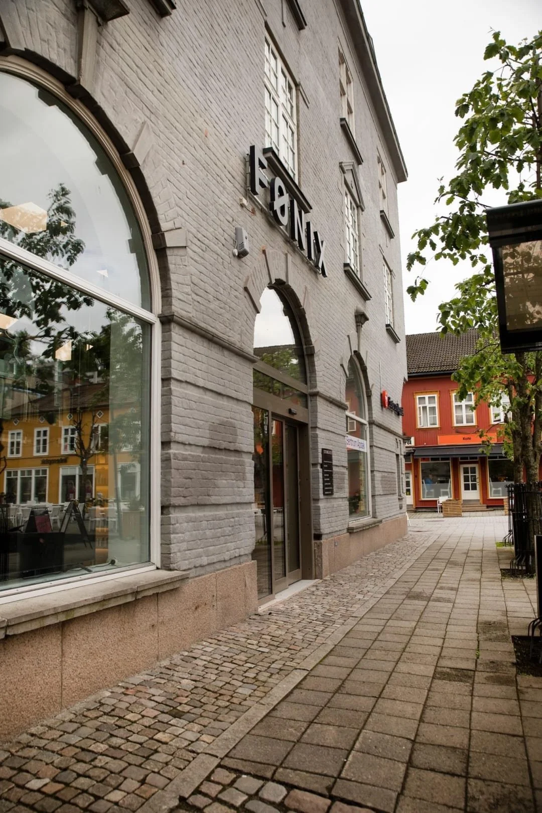 Utsikt av en grå bygning med store vinduer og inngangsdør, omgitt av en koselig gate med brostein og trær, i en liten by.