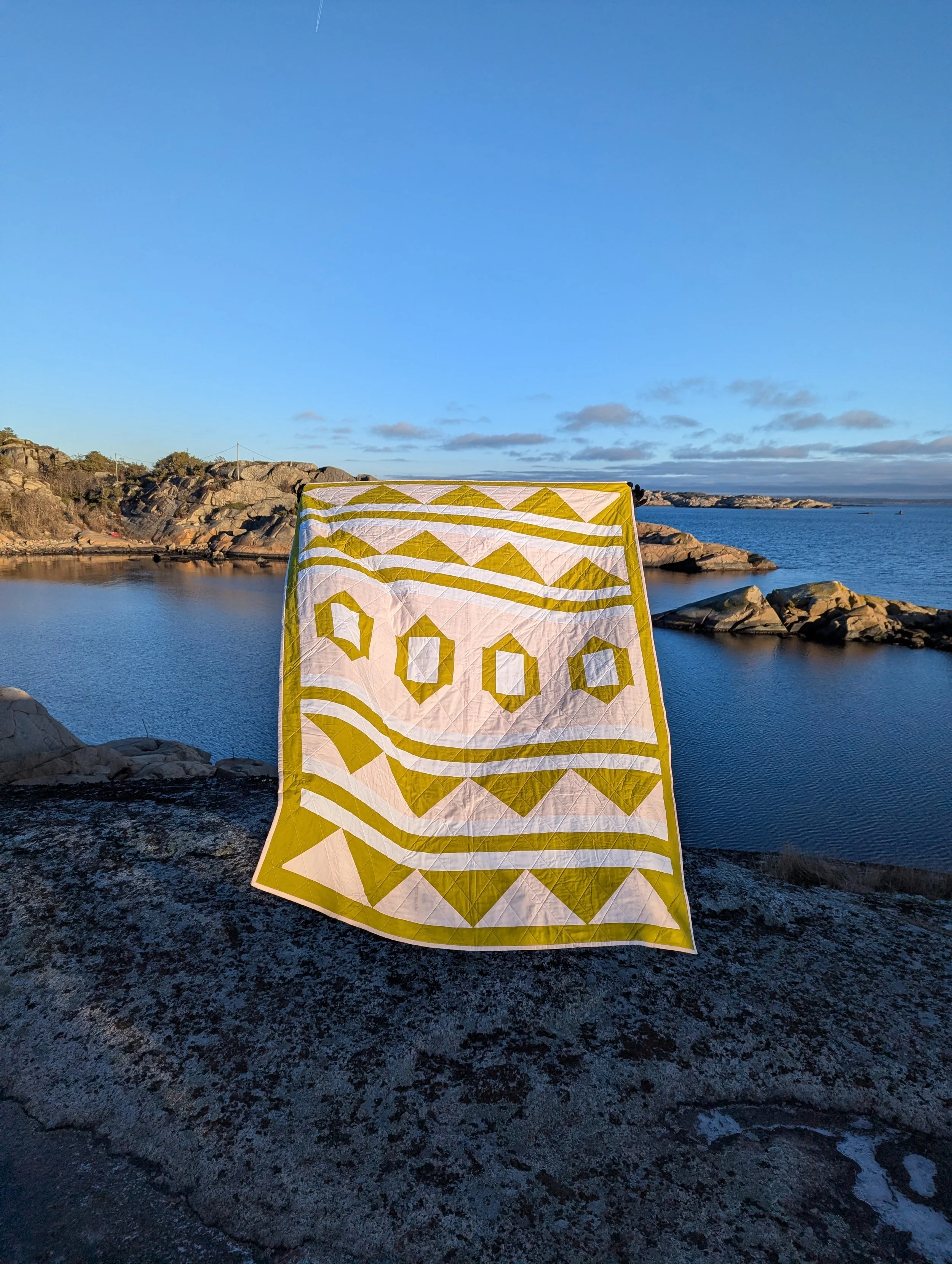 The 2995 Quilt by Millie & Bun Designs- a modern geometric quilt in pink, yellow, and white being held with Norwegian landscape in the background