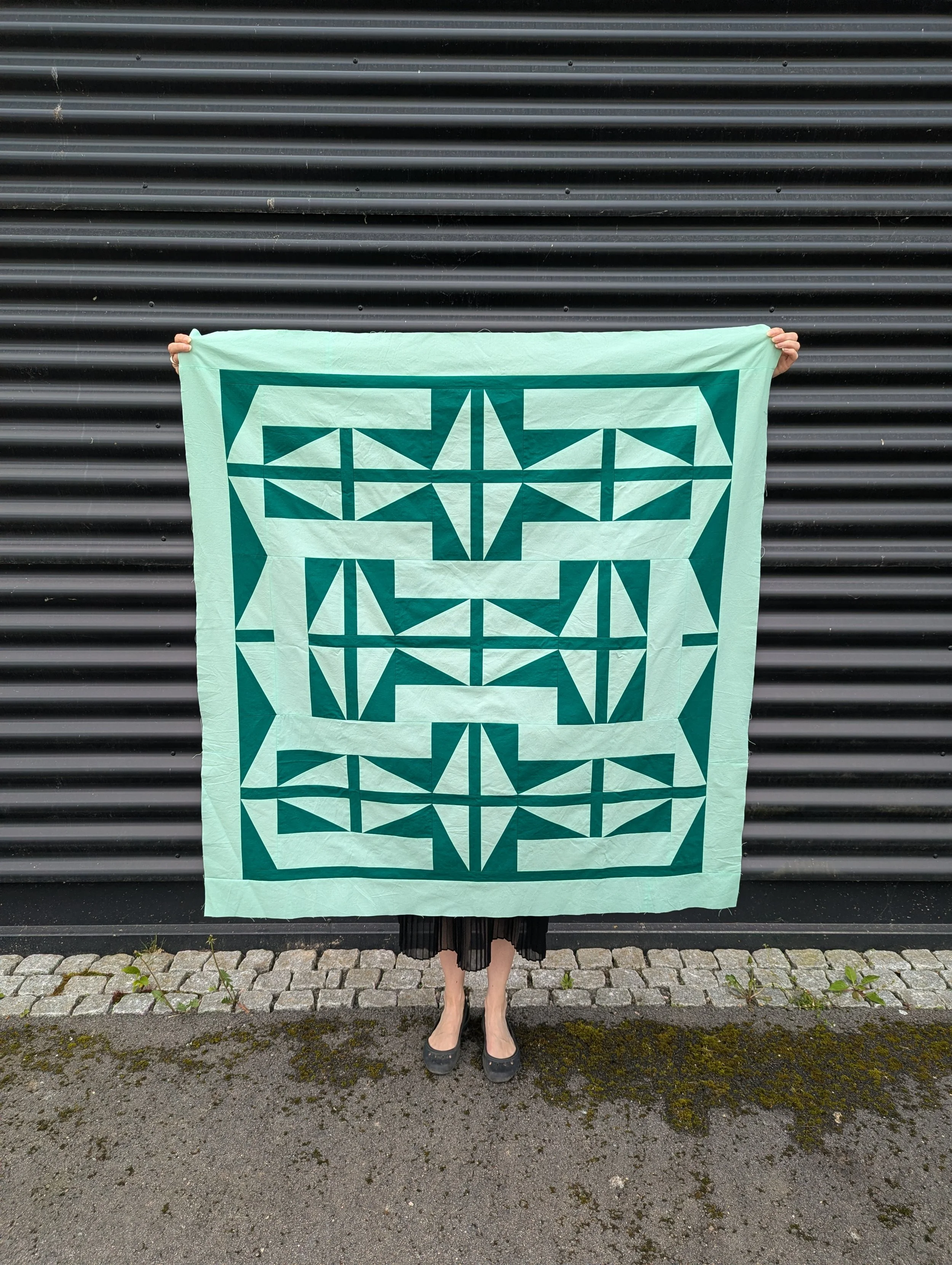 Modern geometric quilt pattern in green colors in front of a black wall