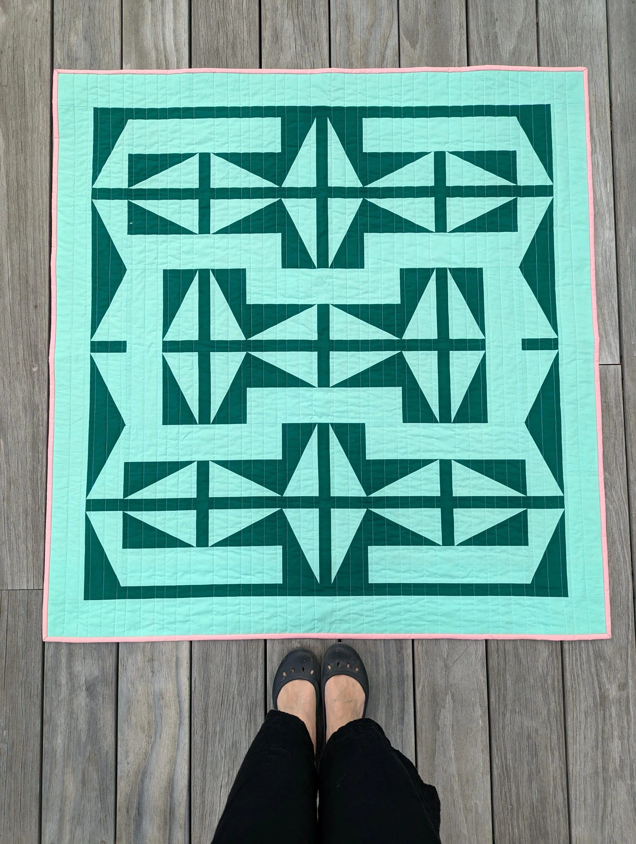 A modern geometric quilt in shades of green with a pop of pink, photographed on a wooden deck with shoes in the frame