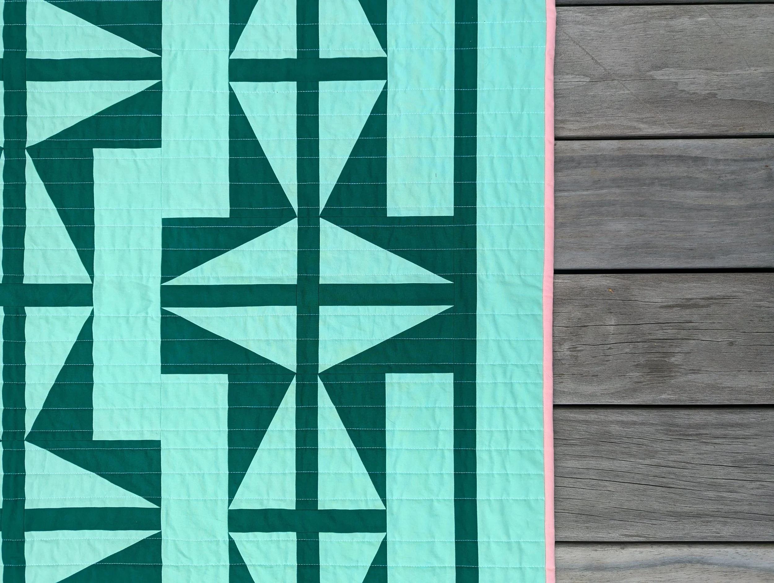 Detail photo of The 1751 Quilt by Millie & Bun Designs- a modern geometric quilt in light and dark green fabric and a pop of pink, with a wooden deck background