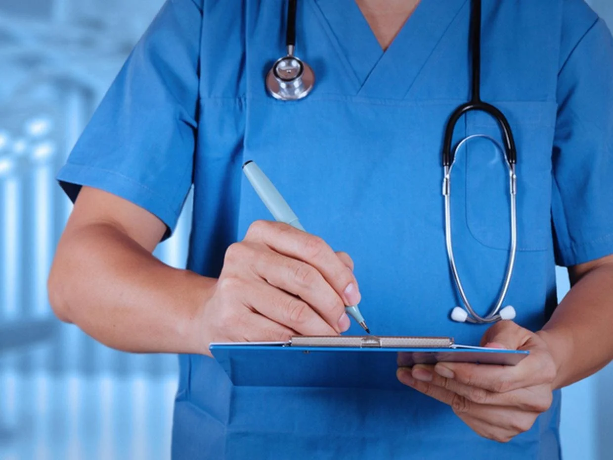 A healthcare professional wearing blue scrubs and a stethoscope, writing on a clipboard with a pen in a hospital or clinical setting.