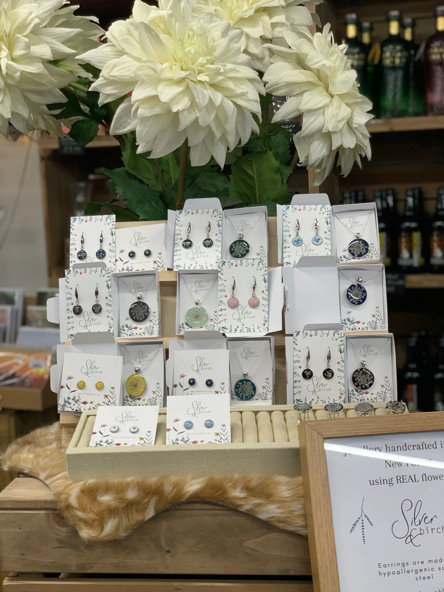 Display of earrings on a wooden stand with large white flowers and a framed sign in the background, showcasing various designs from Silver Birch.