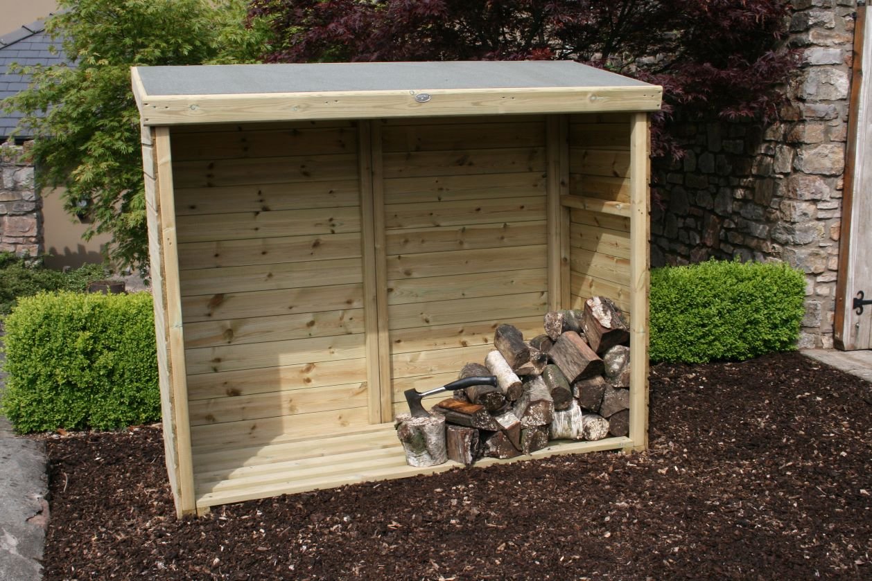 Wood storage shed with firewood and an axe on a gravel area near shrubs and a stone wall.