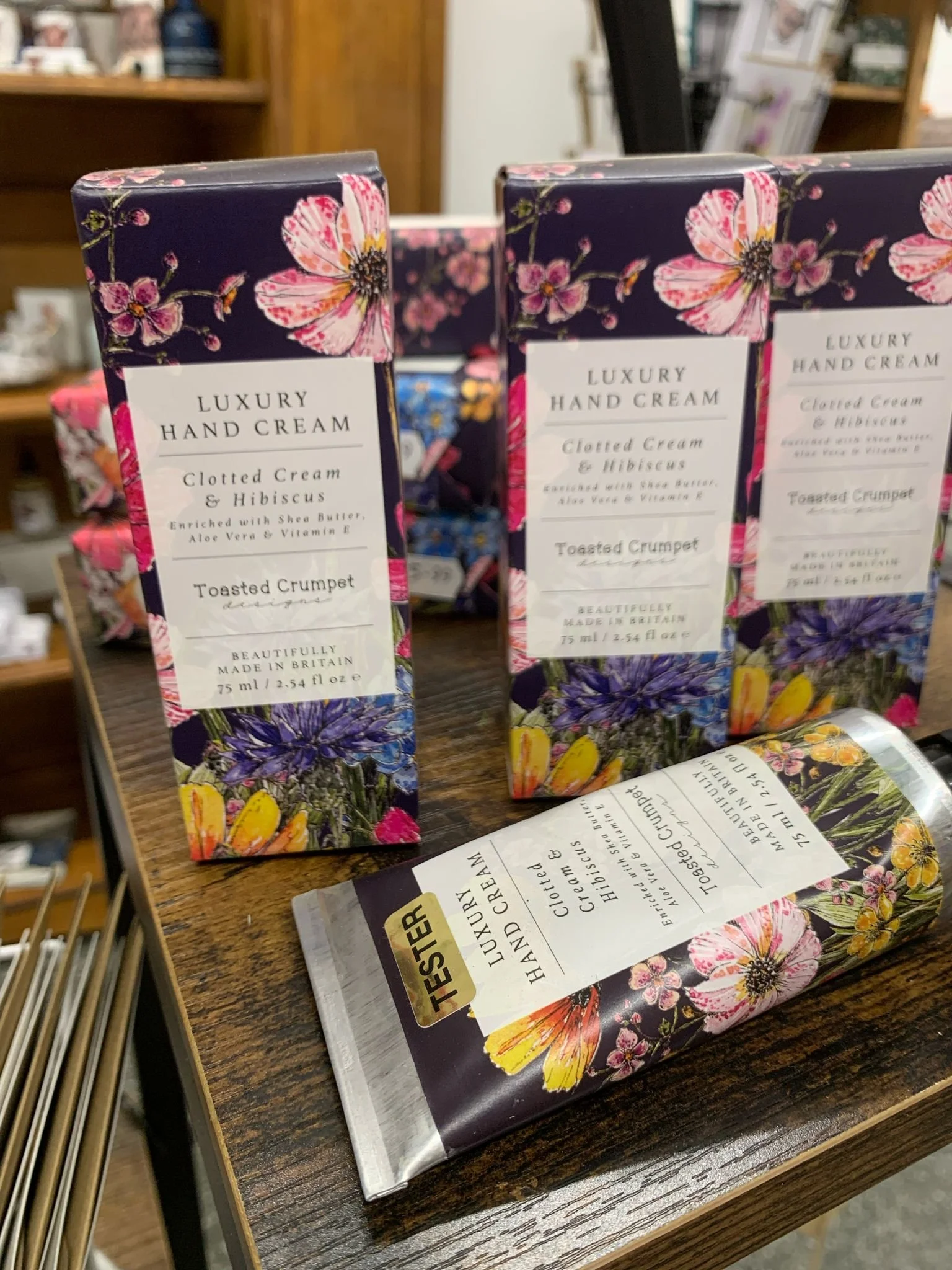 Luxury hand cream display with floral packaging, featuring 'Clotted Cream & Hibiscus' scent by Toasted Crumpet, on a wooden shelf. Tester tube in foreground.