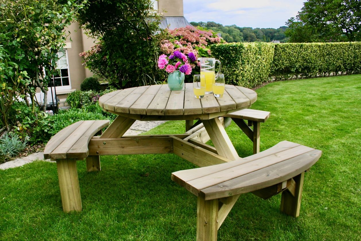 Round wooden picnic table with benches on green lawn, featuring a vase of flowers and glasses of orange juice.