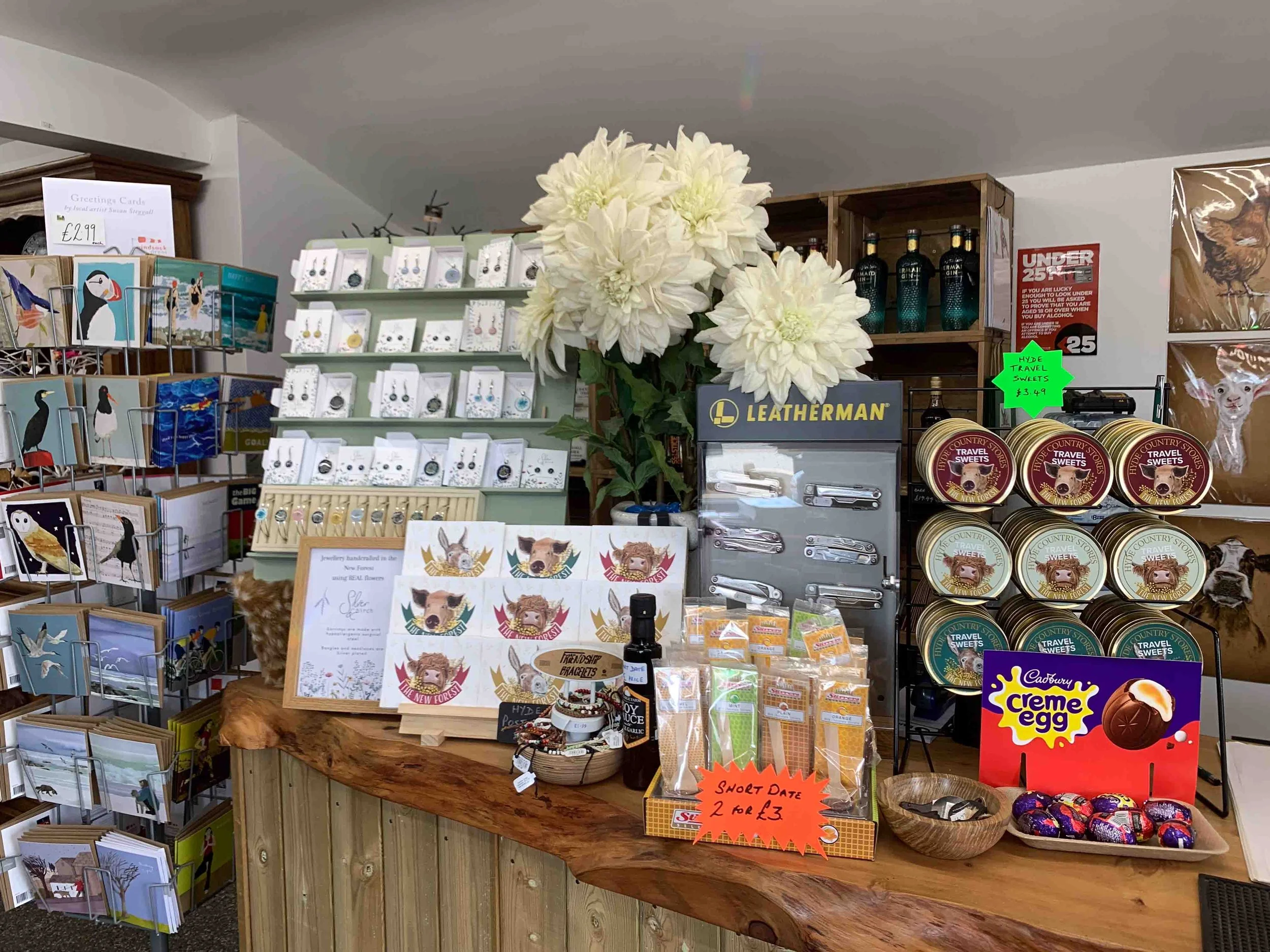 Store display featuring cards with animal designs, handmade jewelry, sweet treats including Cadbury Creme Eggs, and Leatherman tools, with decorative white flowers on a wooden counter.