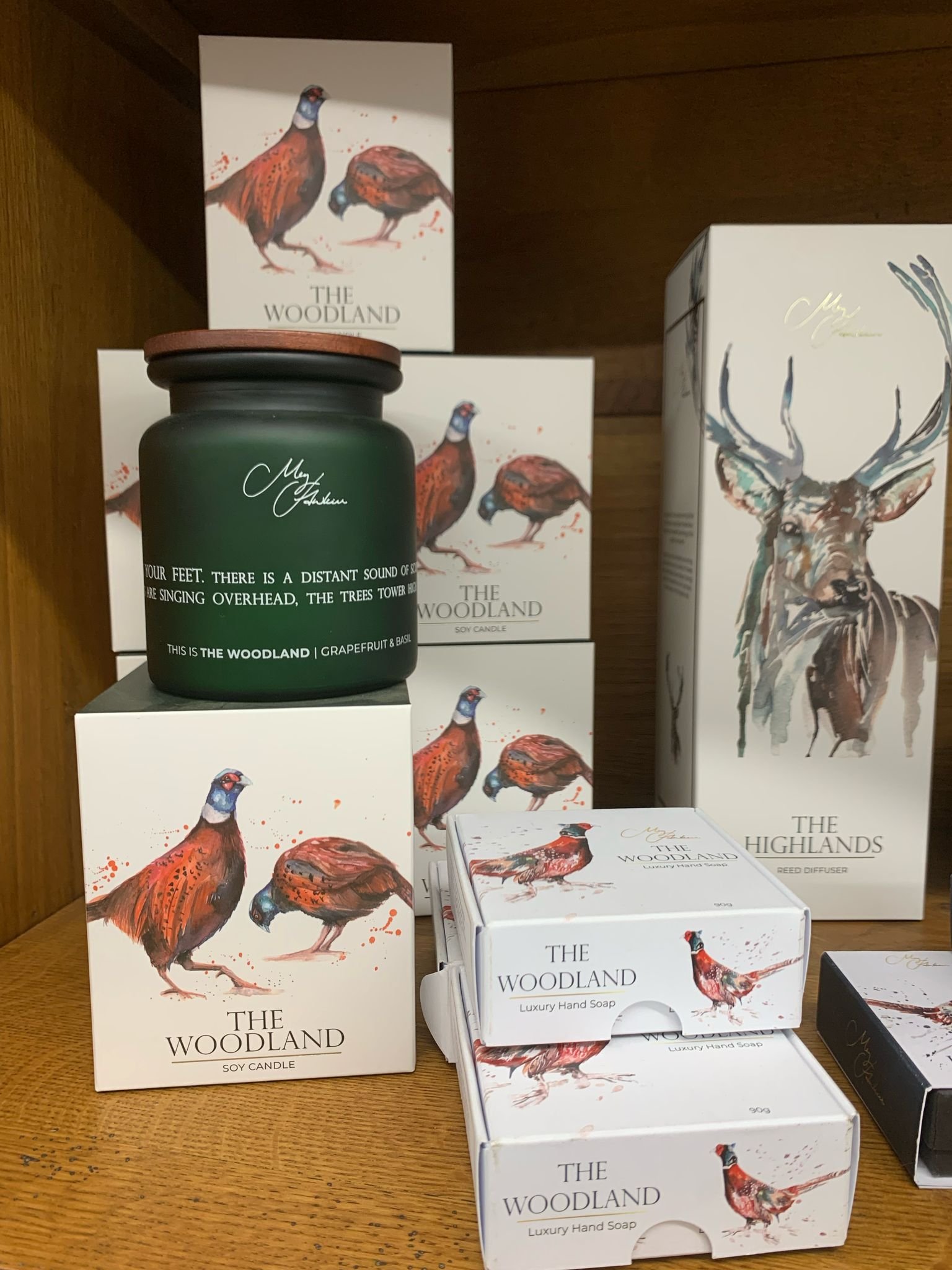 Display of "The Woodland" scented products featuring soy candles and luxury hand soap with packaging depicting pheasants. A "The Highlands" reed diffuser box with stag illustration is also visible. Products are on a wooden shelf.
