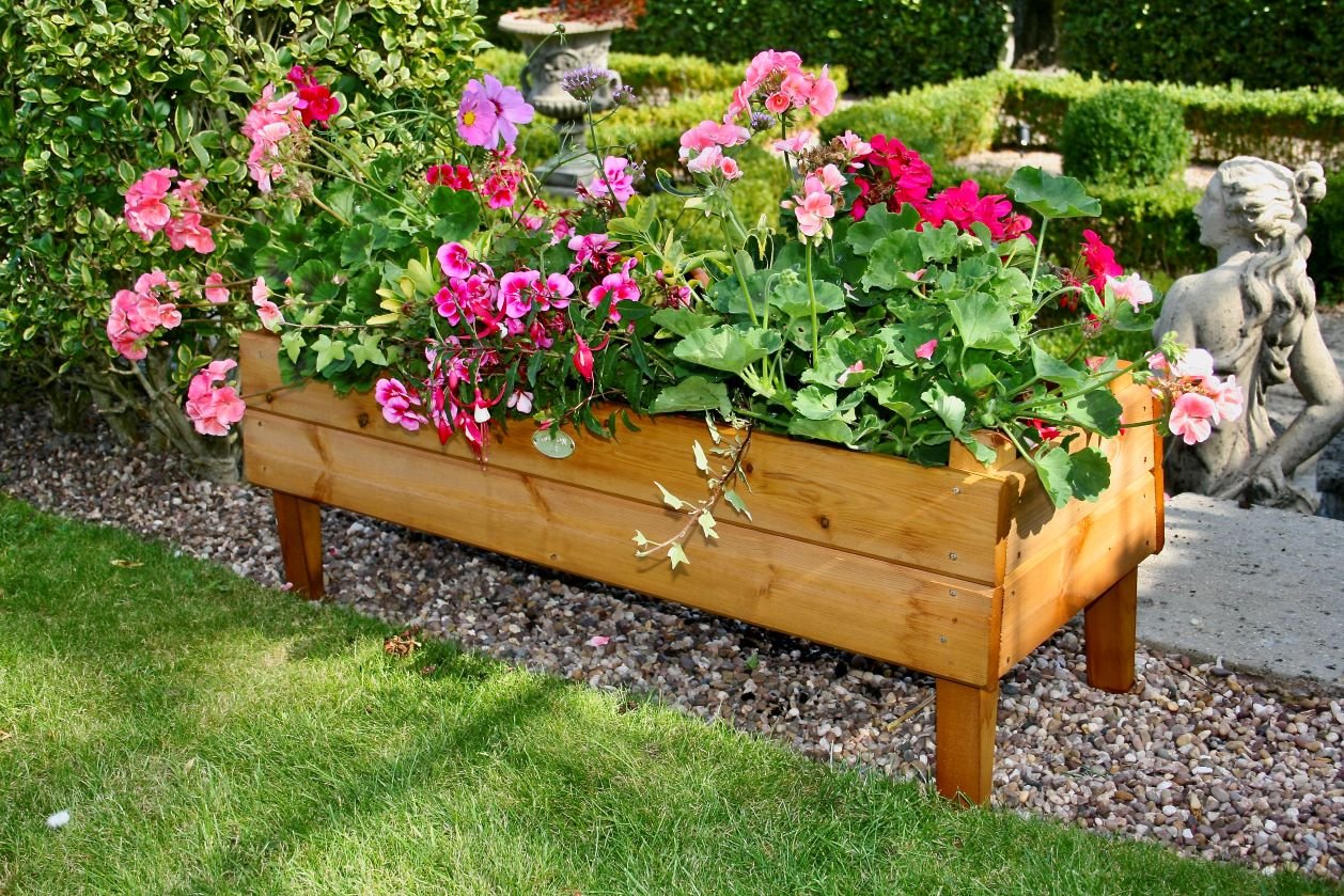 Wooden planter box with colorful flowers in a garden setting, surrounded by hedges and a stone statue.