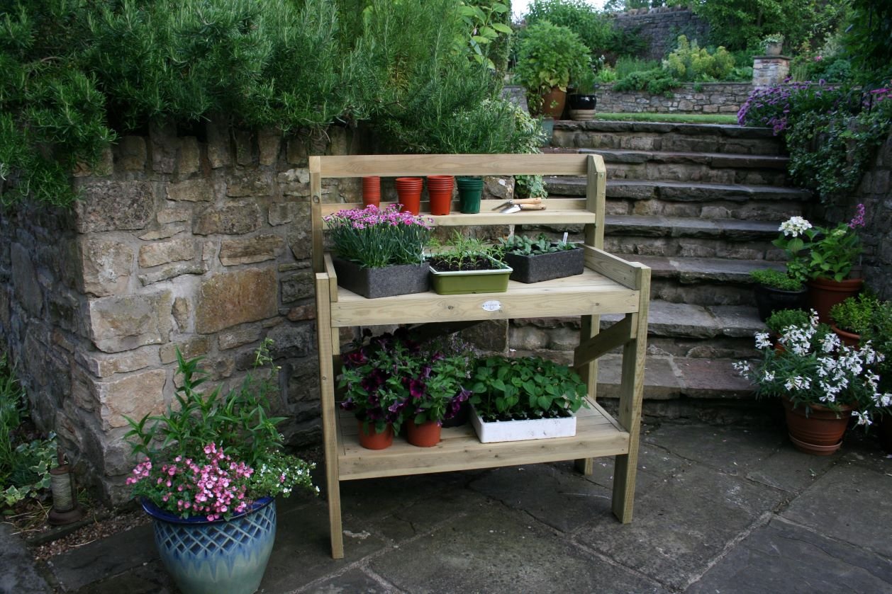 Wooden potting bench with potted plants and gardening tools in a garden with stone steps and greenery.