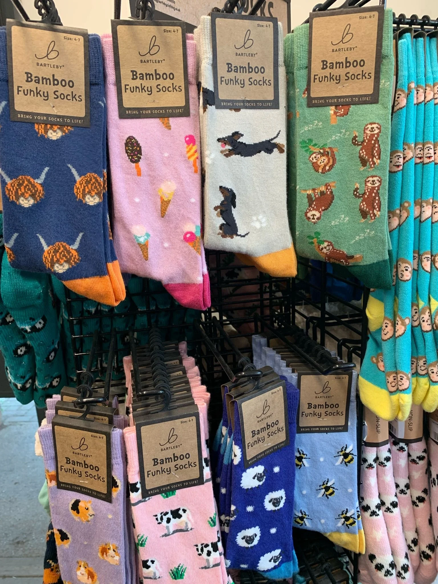 Display of colorful bamboo socks with various animal and food patterns, including tigers, ice creams, dogs, sloths, and bees, labeled "Bartleby Bamboo Funky Socks."