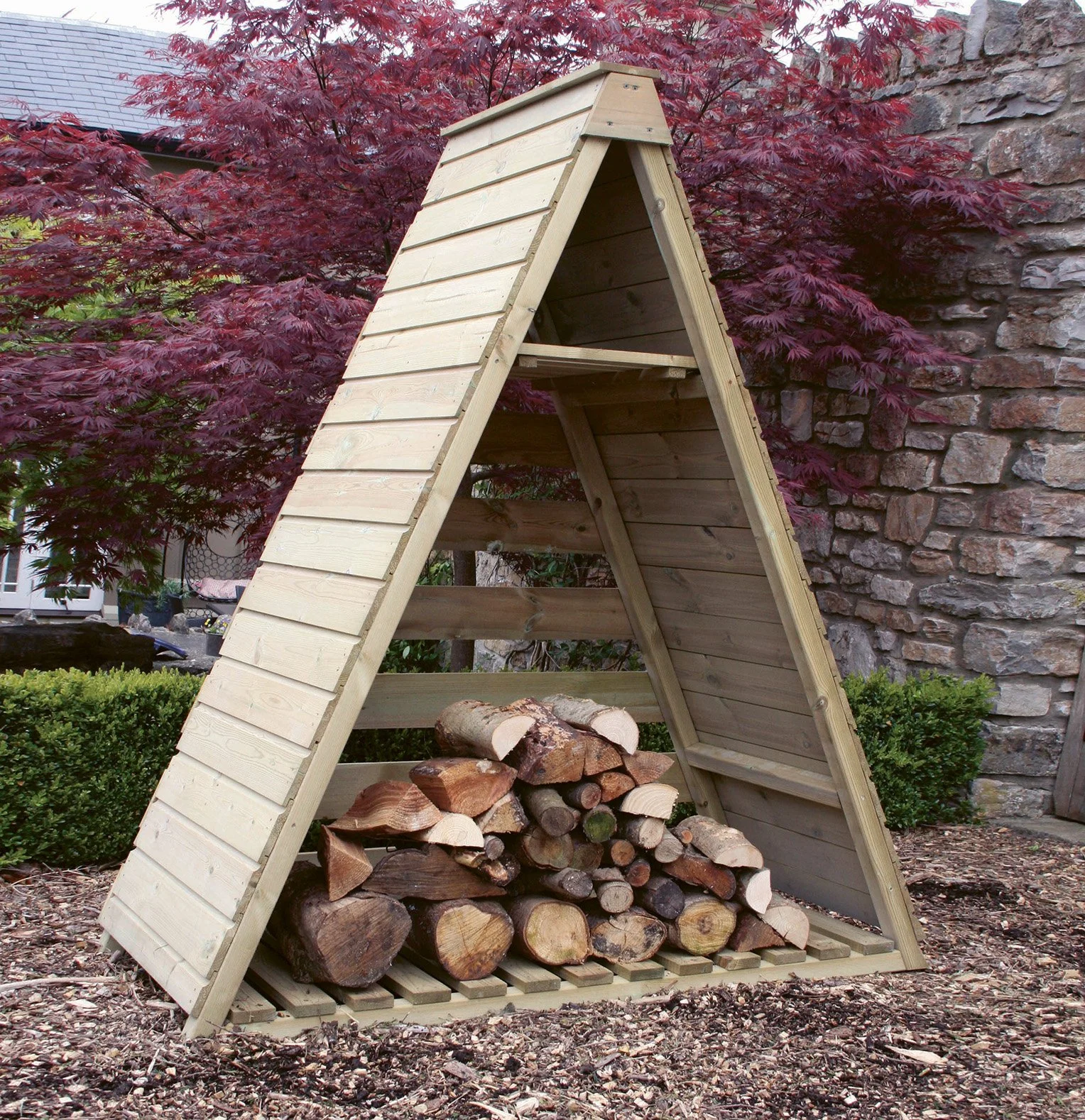 A-frame wooden log store filled with firewood outdoors against a stone wall, with a tree in the background.