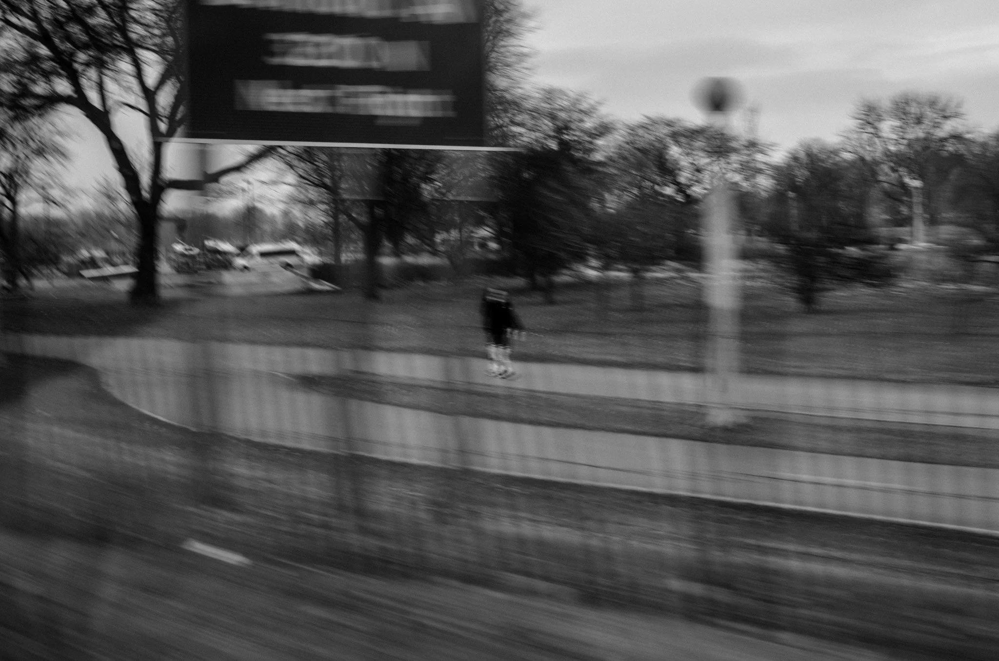 A black and white photo taken from a moving vehicle showing a park with leafless trees, a person walking on the sidewalk, a lamppost, and a blurred street sign in the background.