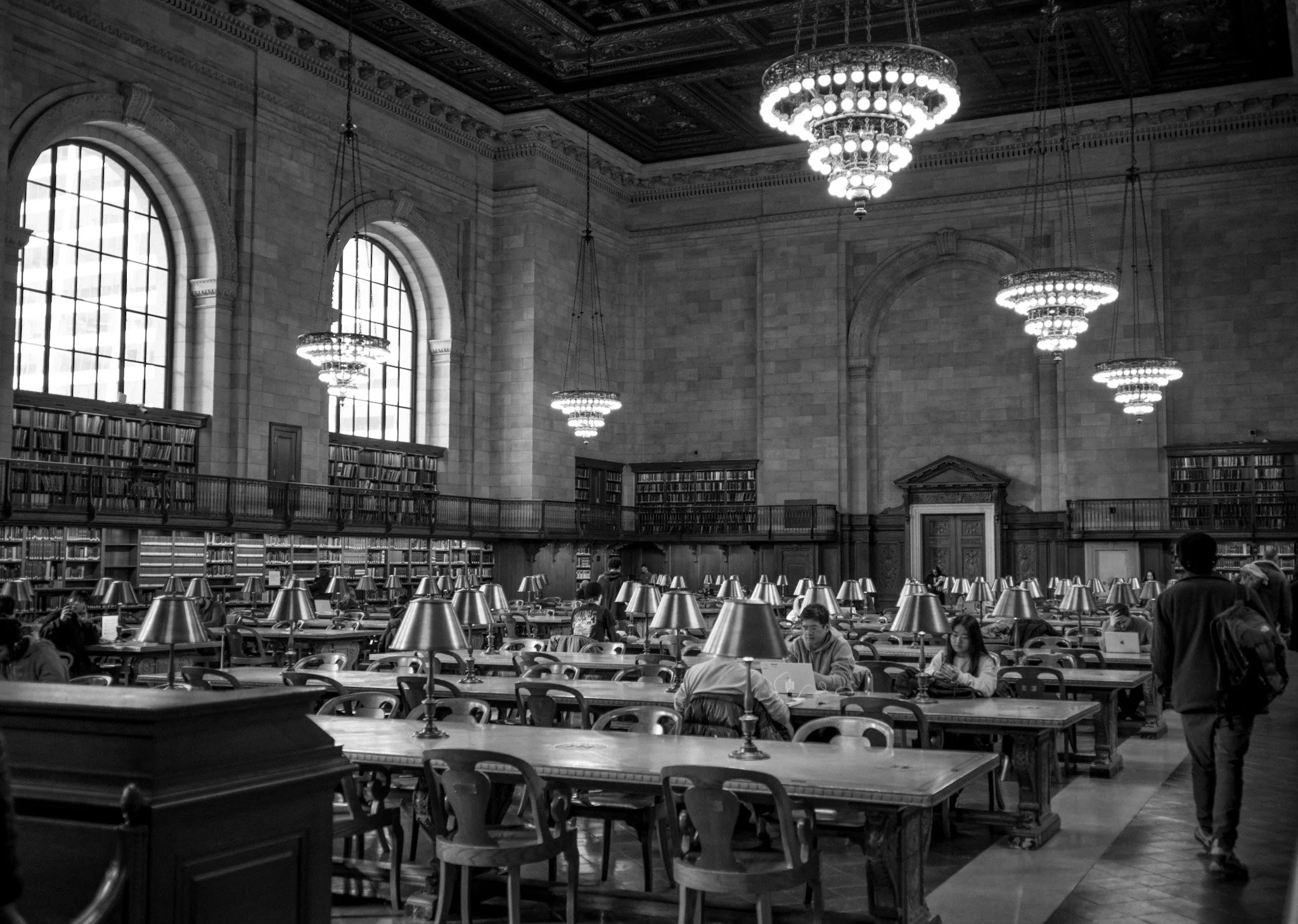 Interior of a grand library hall with high arched windows, ornate ceiling, chandeliers, and multiple rows of tables with reading lamps, filled with people reading and studying.