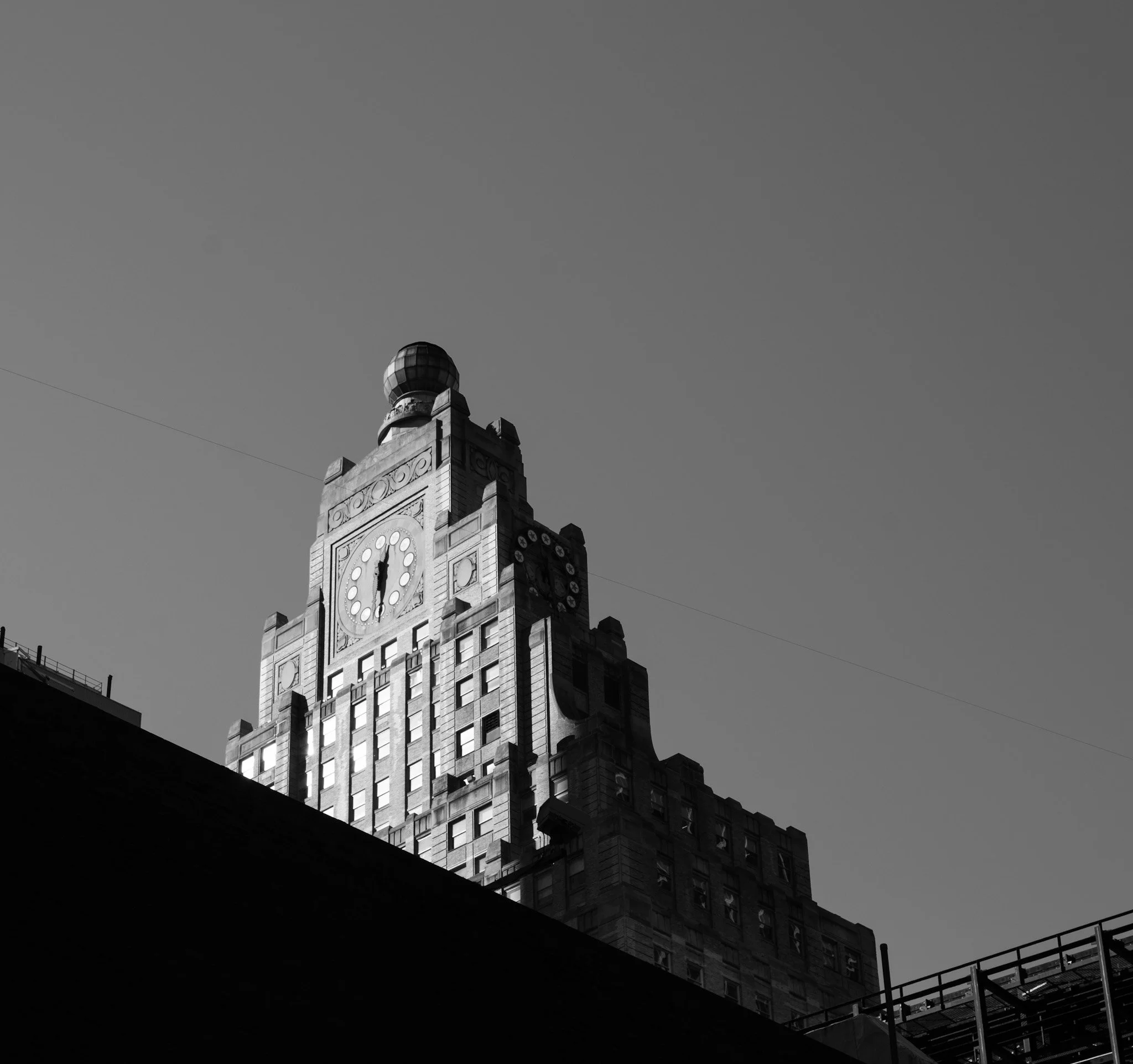 Black and white photo of a clock tower building with a rounded top, ornate architectural details, a clock face showing 12:02, and a dark shadow in the foreground under a clear sky.