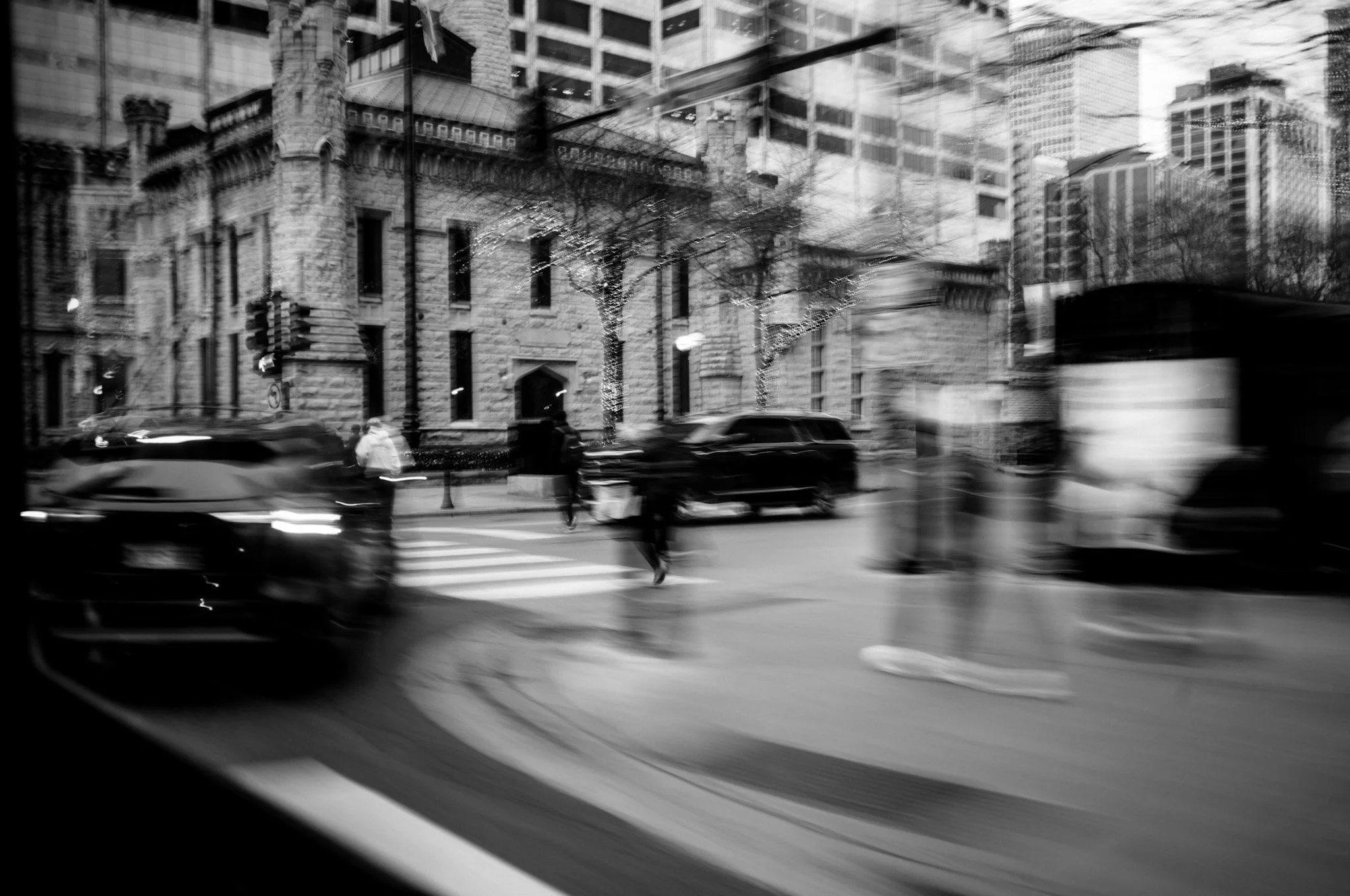 Black and white photo of city streets with cars and pedestrians, blurred motion effect, tall buildings, trees with lights, and traffic signals.