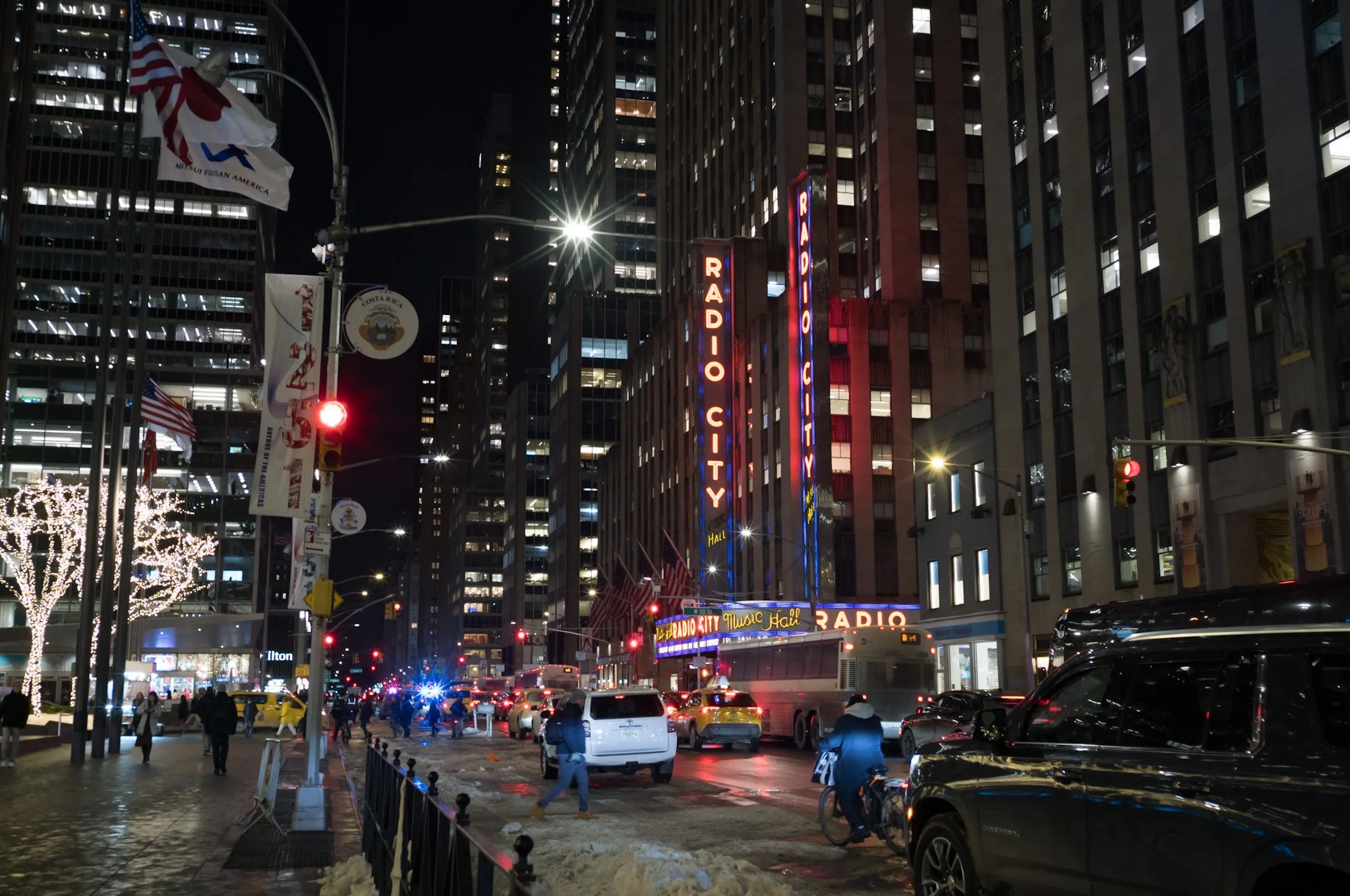 Nighttime city street scene with tall buildings, neon signs, and crowd of pedestrians walking on sidewalk. Cars parked along street, some moving, with bright streetlights illuminating scene.