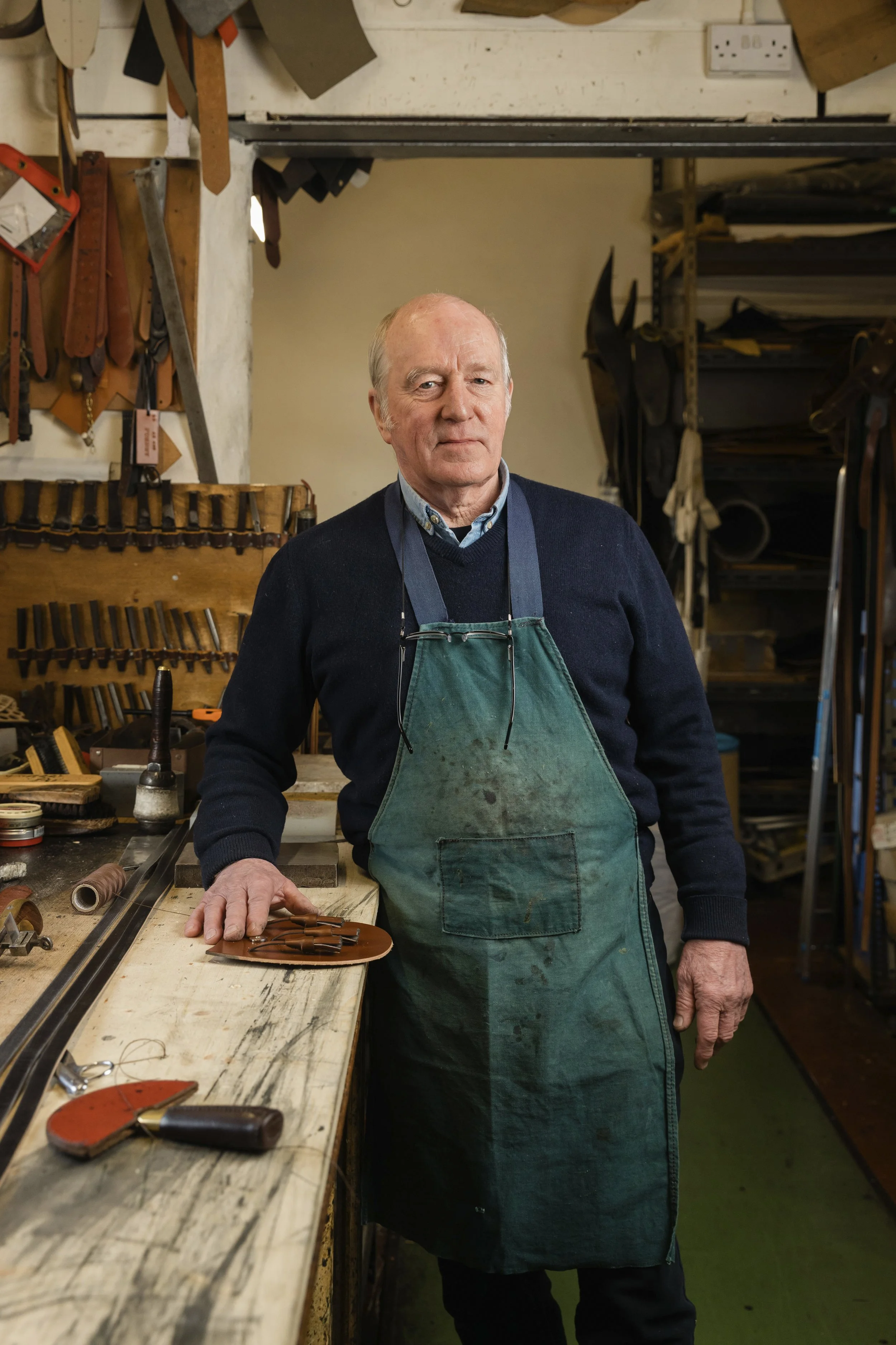 An elderly man standing in a woodworking workshop, wearing a green apron and surrounded by tools and woodworking supplies.