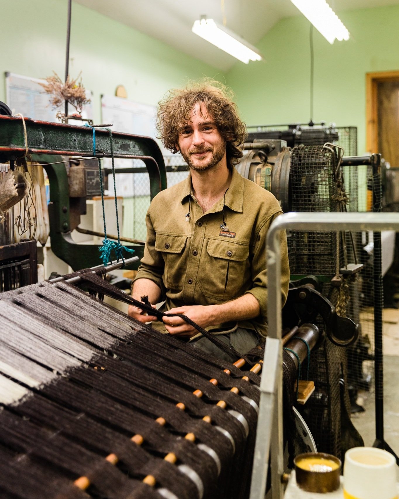 A man with curly hair and a beard working on a loom in a workshop with green walls and fluorescent lighting. Documentary Photography by Mark Gillies. 