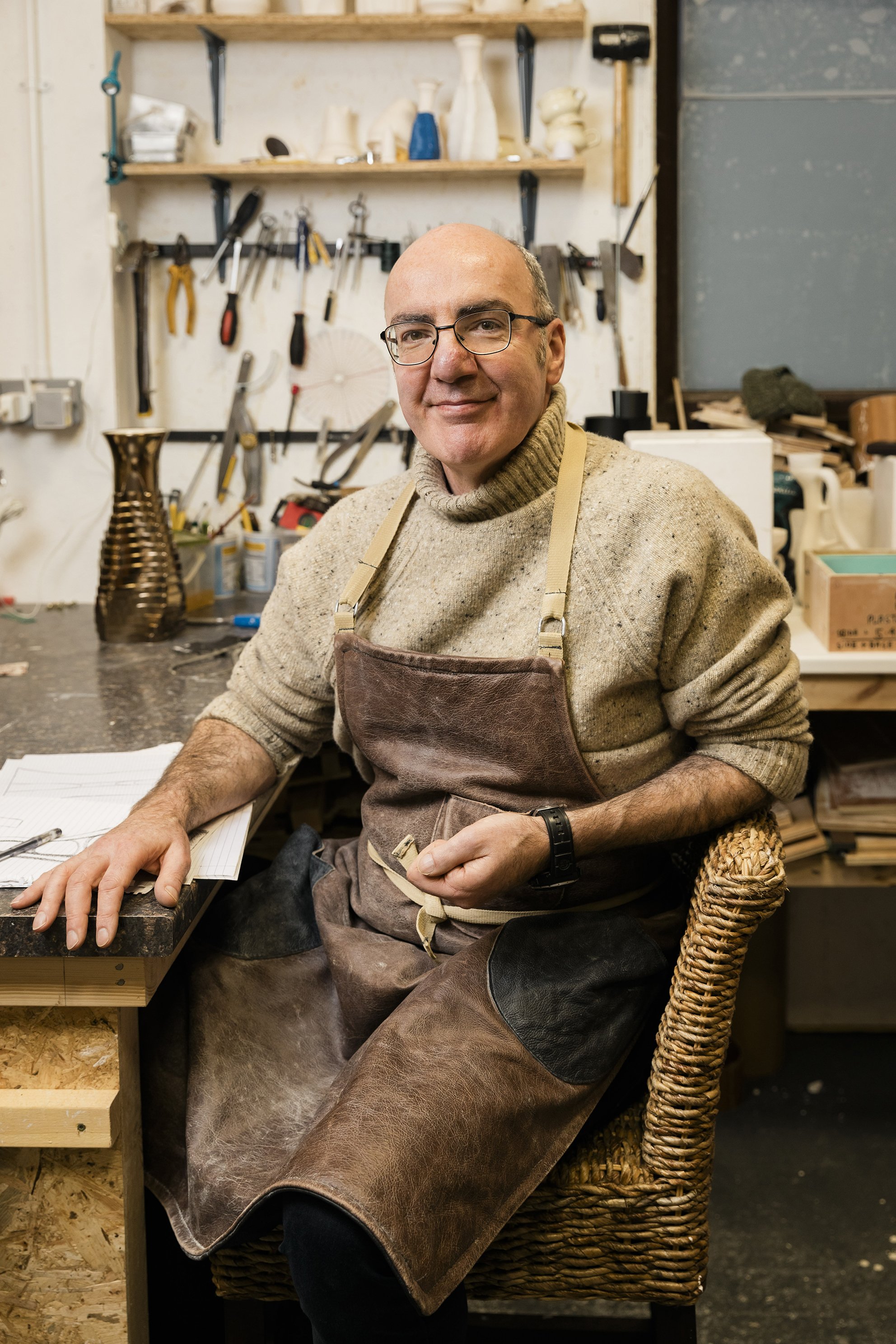 A man in a workshop, wearing glasses and a beige sweater, sitting on a woven chair at a workbench with tools and materials, surrounded by shelves with pottery and woodworking tools. Documentary portrait shot by Mark Gillies. 