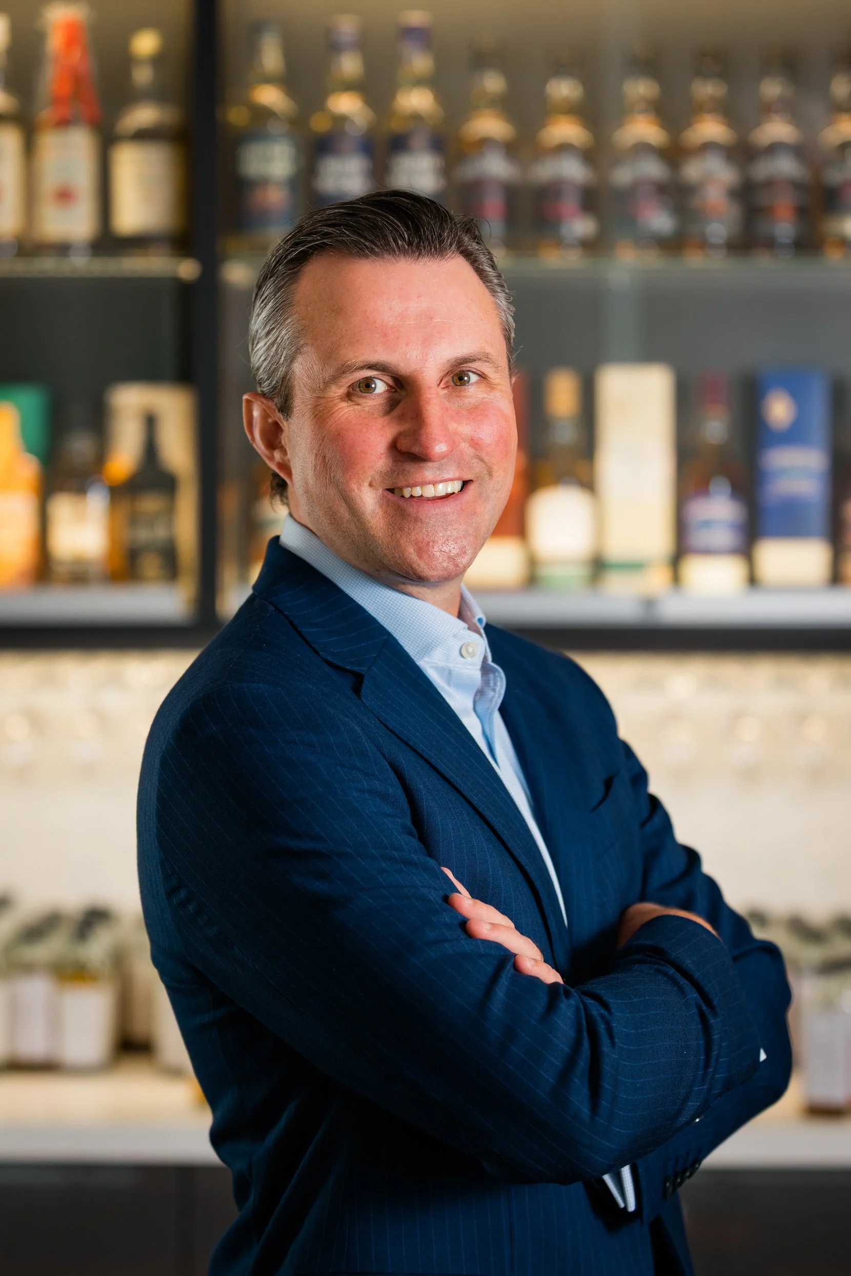 A man in a blue suit with arms crossed, smiling in front of shelves of bottles. Corporate headshot portrait photography shot by Mark Gillies based in Glasgow. This image features CEO of Whyte and McKay Whisky.  