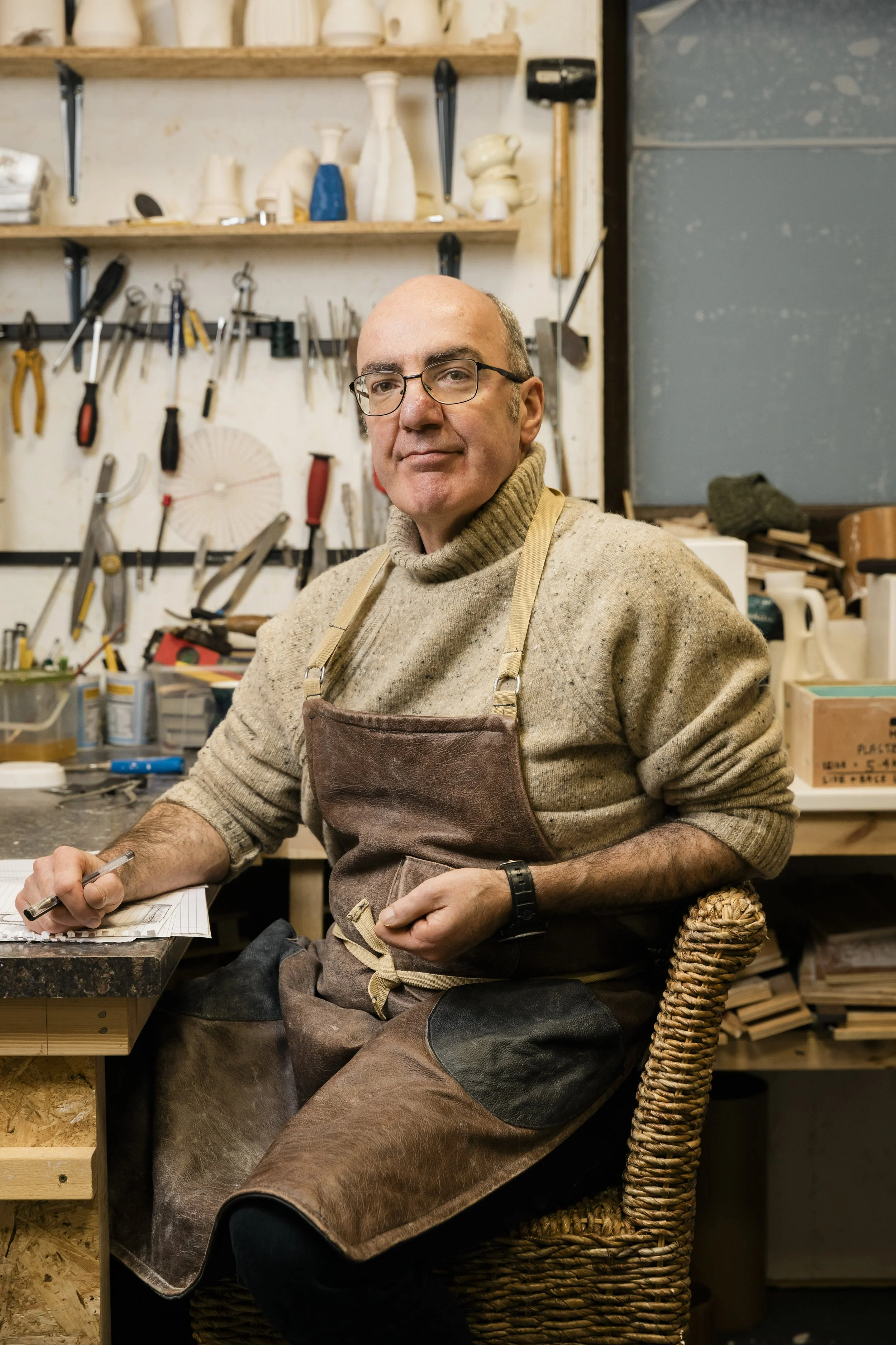 A man in a workshop, wearing glasses and a beige sweater, sitting on a woven chair at a workbench with tools and materials, surrounded by shelves with pottery and woodworking tools. Documentary portrait shot by Mark Gillies. 