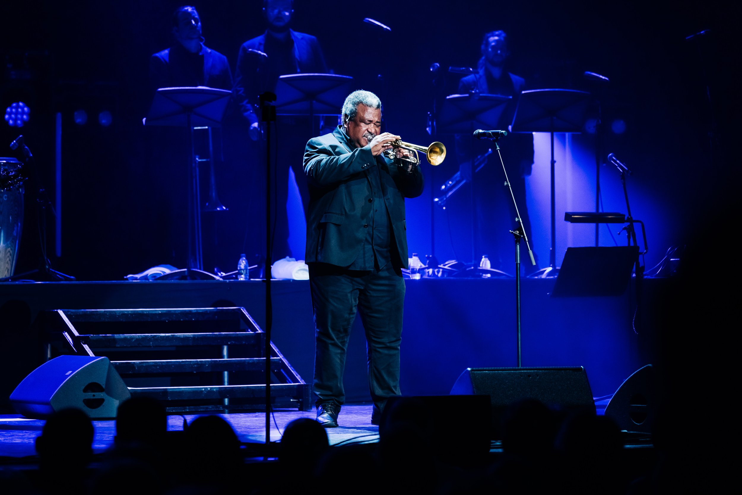 A man playing trumpet on stage illuminated by blue light, with a music band in the background, and audience silhouettes in the foreground. Live event concert photography at Royal Concert Hall in Glasgow by Mark Gillies for Buena Vida All Stars. 