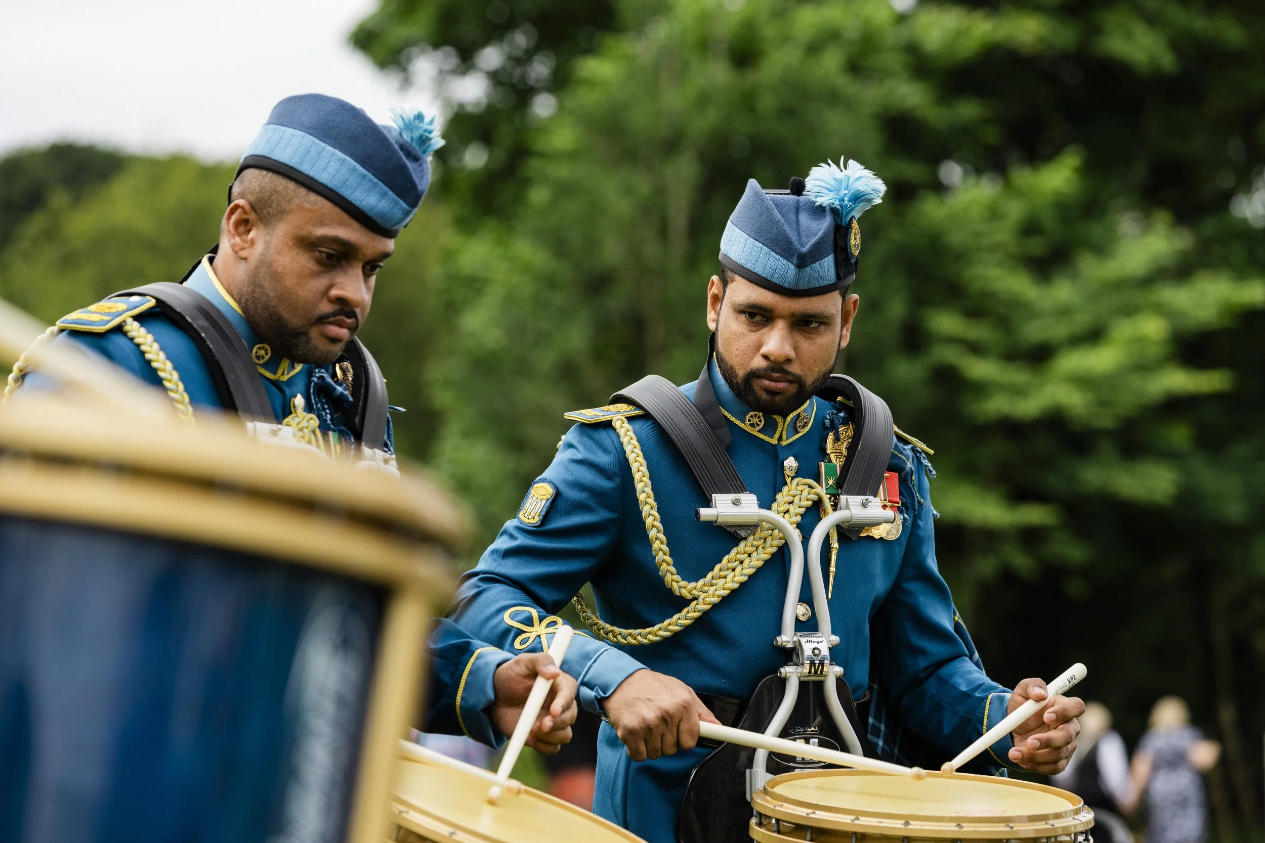 Two uniformed musicians playing drums outdoors, wearing blue military-style uniforms with medals and gold cords, and blue hats with feathers. Reportage photography by Mark Gillies Photography based in Glasgow for the Dundonald Highland Games. 