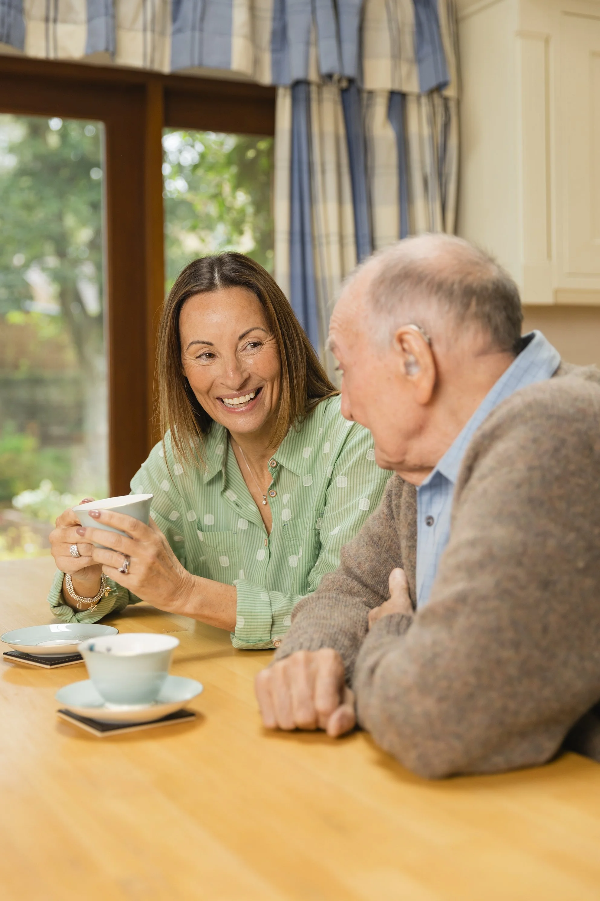 A woman and an elderly man sitting at a table, enjoying a conversation while holding cups. Commercial photography shot by Mark Gillies, stock imagery created for Carents charity. 