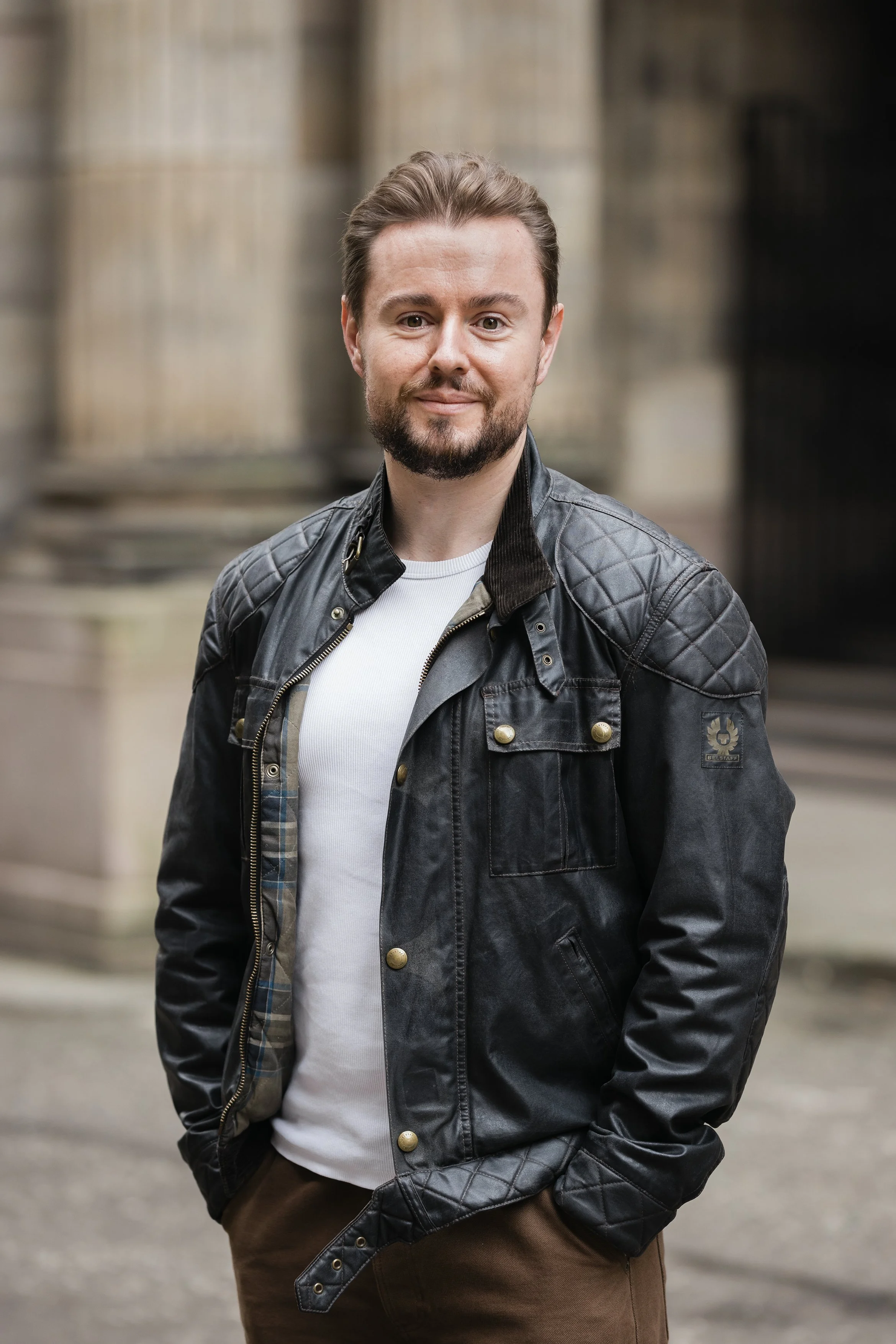 Portrait of a young man with beard, wearing a black leather jacket and white shirt, standing outdoors with blurred stone columns in the background. Headshot portrait photography shot by Mark Gillies based in Glasgow. 