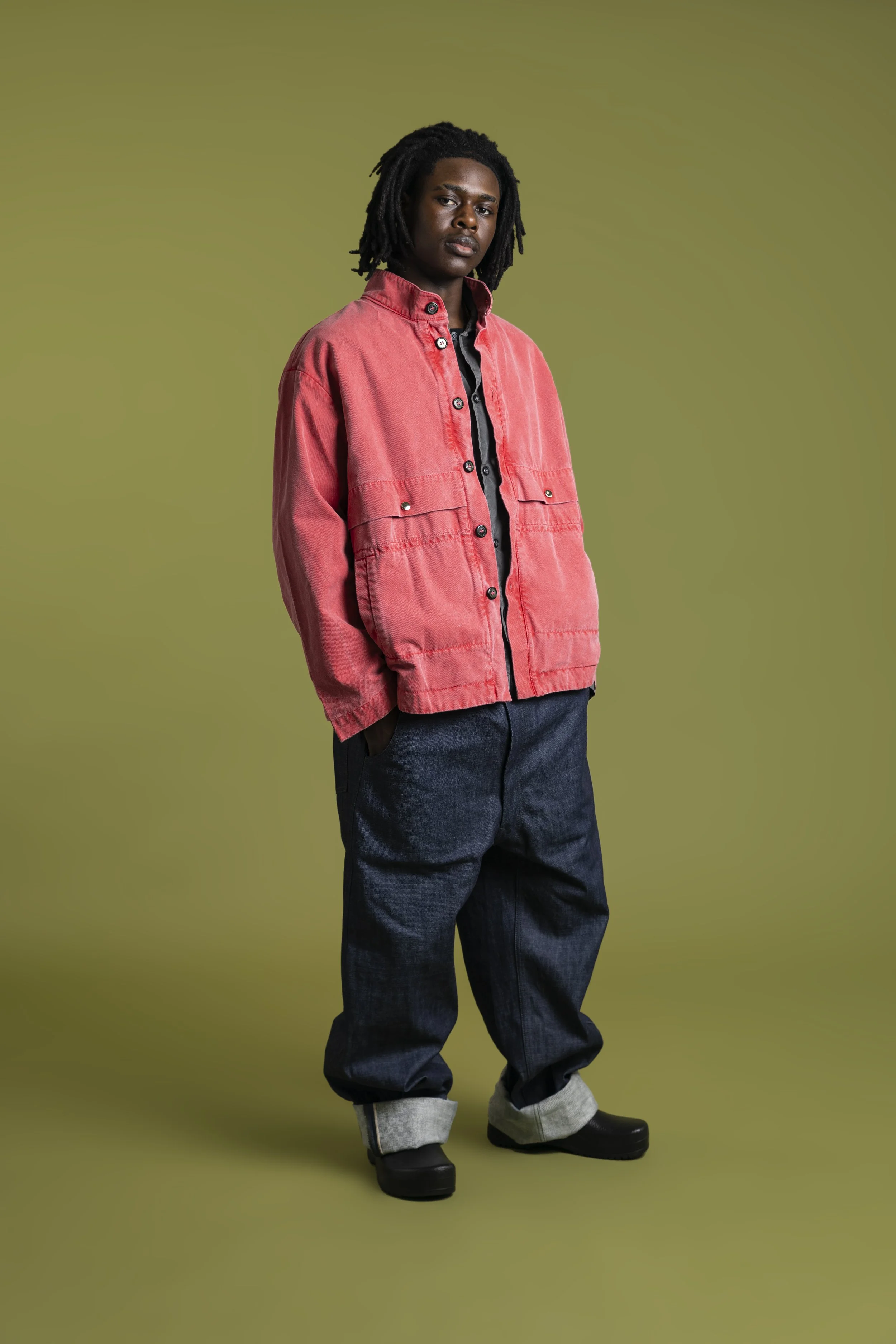 A young man with dreadlocks wearing a red jacket, black shirt, dark jeans, and black shoes standing against a plain green background. Fashion photography shot by Mark Gillies Photography. Image shot for Edwin Clothing. 