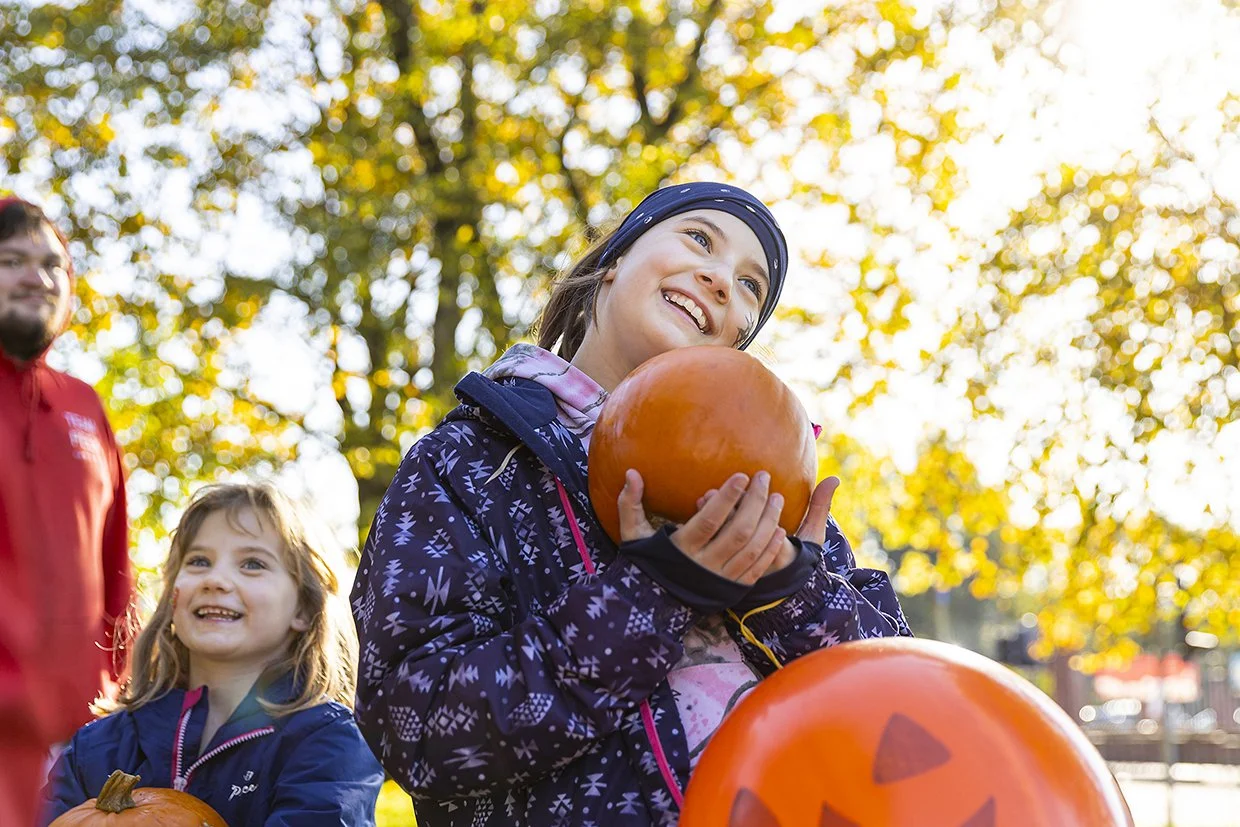 Smiling young girl holding a pumpkin with a carved face, outdoors in fall with trees and sunlight in the background, with other children and adults around.