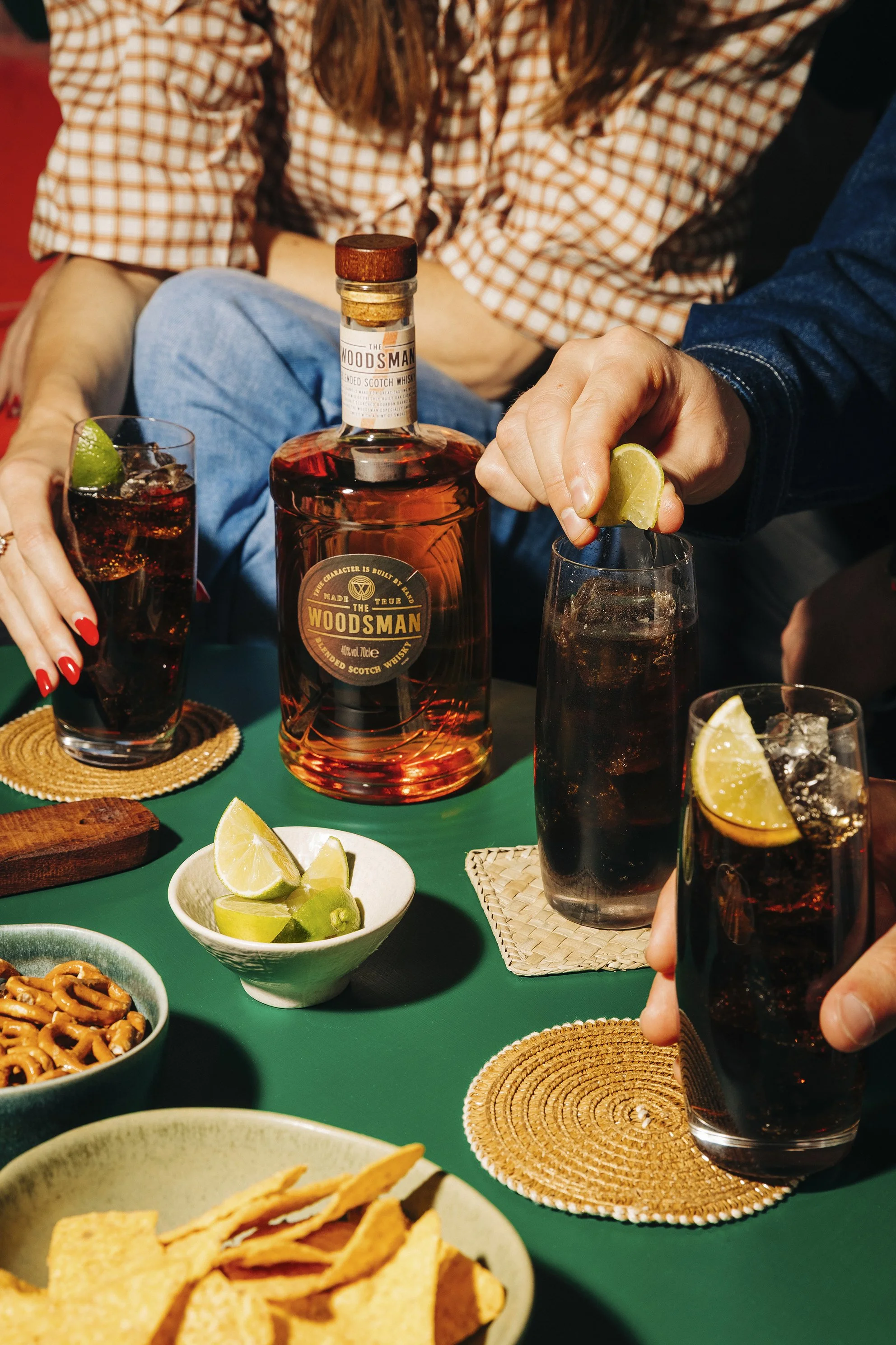 Friends enjoying drinks with whiskey on a green table, lime wedges, snacks like pretzels and chips, and coasters. Commercial whisky photography shot by Mark Gillies Photography based in Glasgow. Image shot for Whyte and McKay Whisky. 