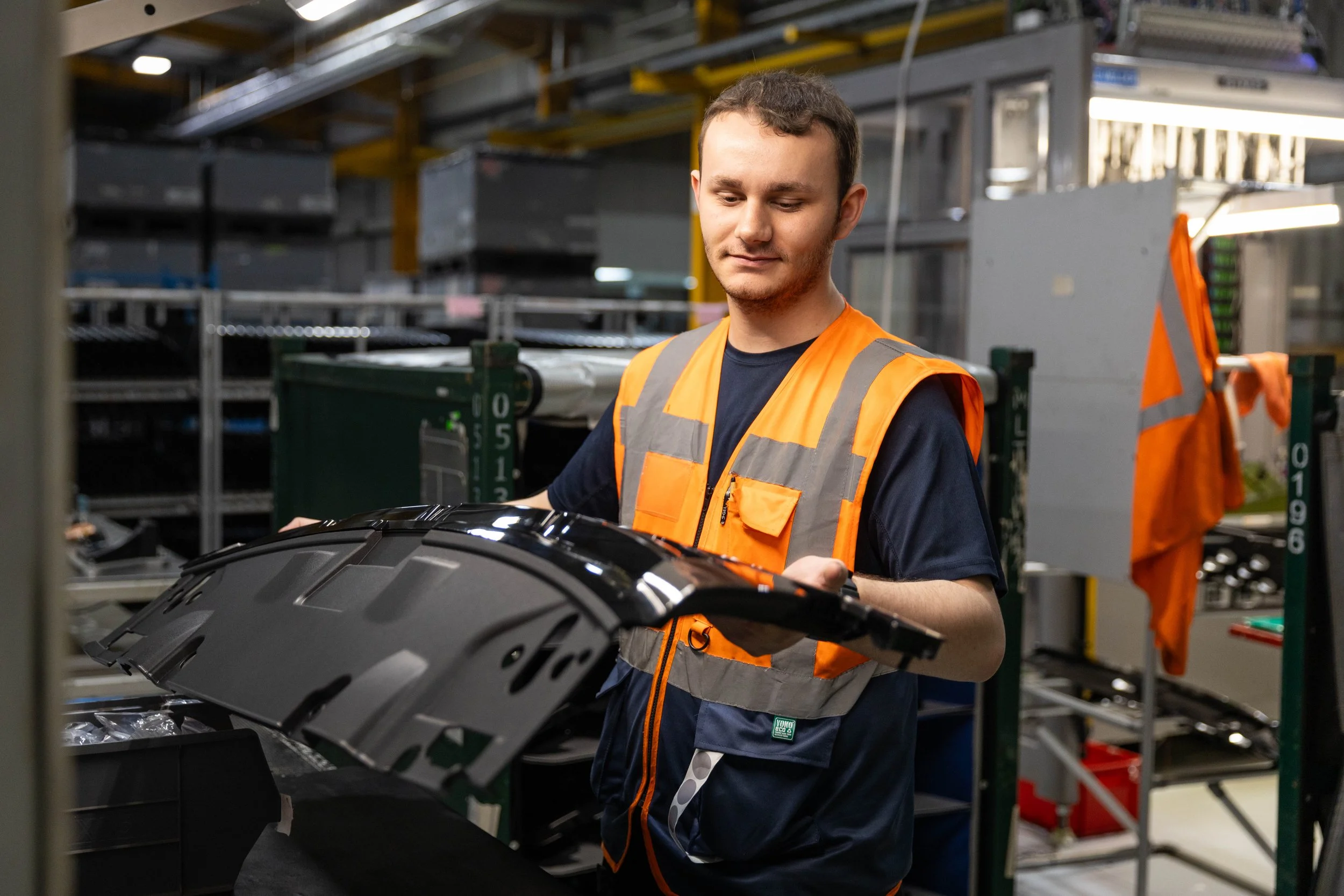 A worker in an orange safety vest examining a black car part in a warehouse. Commercial photography image taken by Mark Gillies.