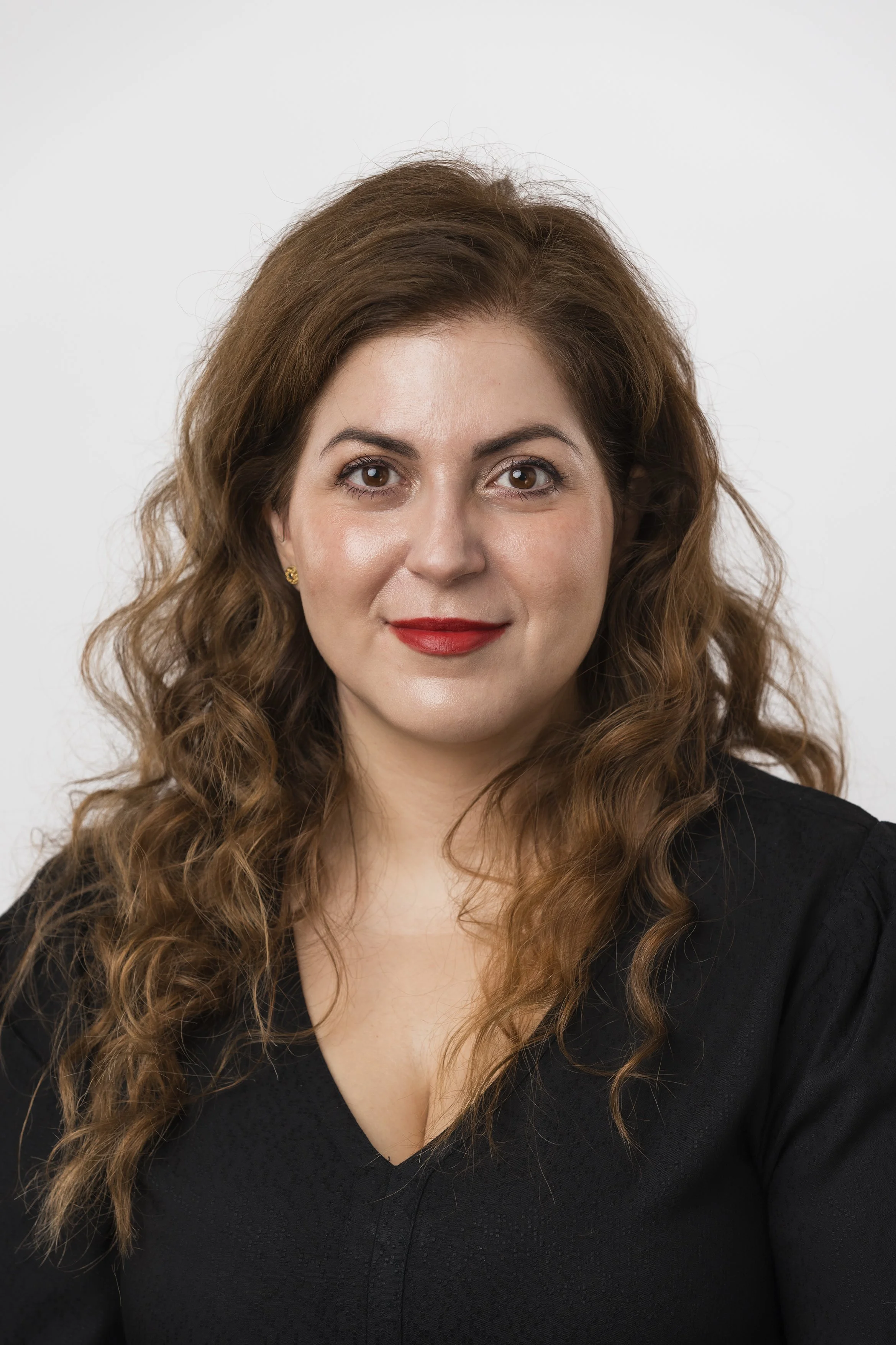 A woman with long, curly brown hair and red lipstick, wearing a black top, smiling softly against a plain white background. Corporate headshot portrait photography shot by Mark Gillies based in Glasgow. Image shot for Klira Skin. 
