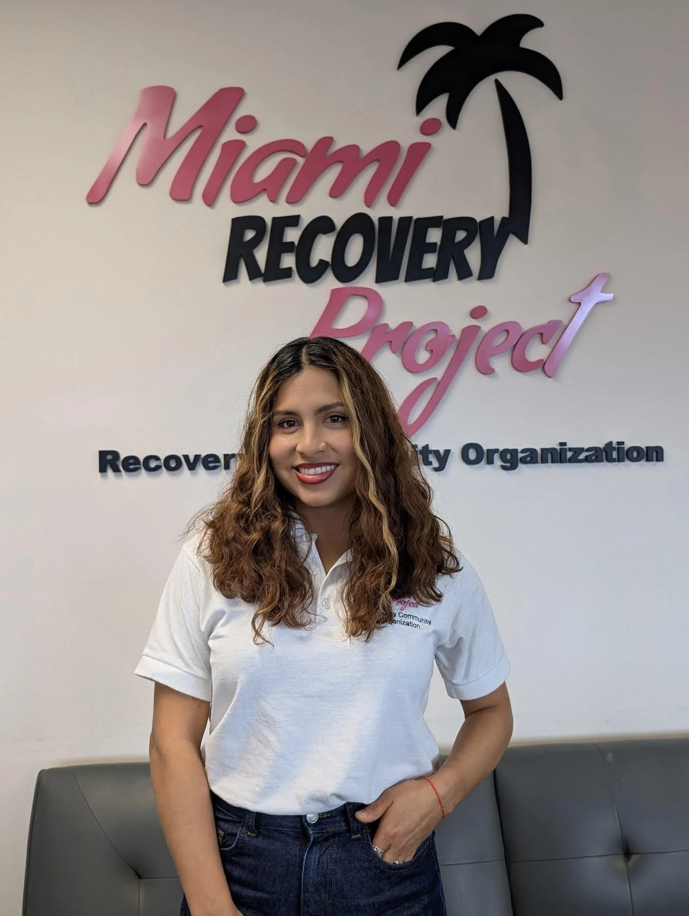 Woman standing in front of Miami Recovery Project sign, wearing a white shirt and jeans.