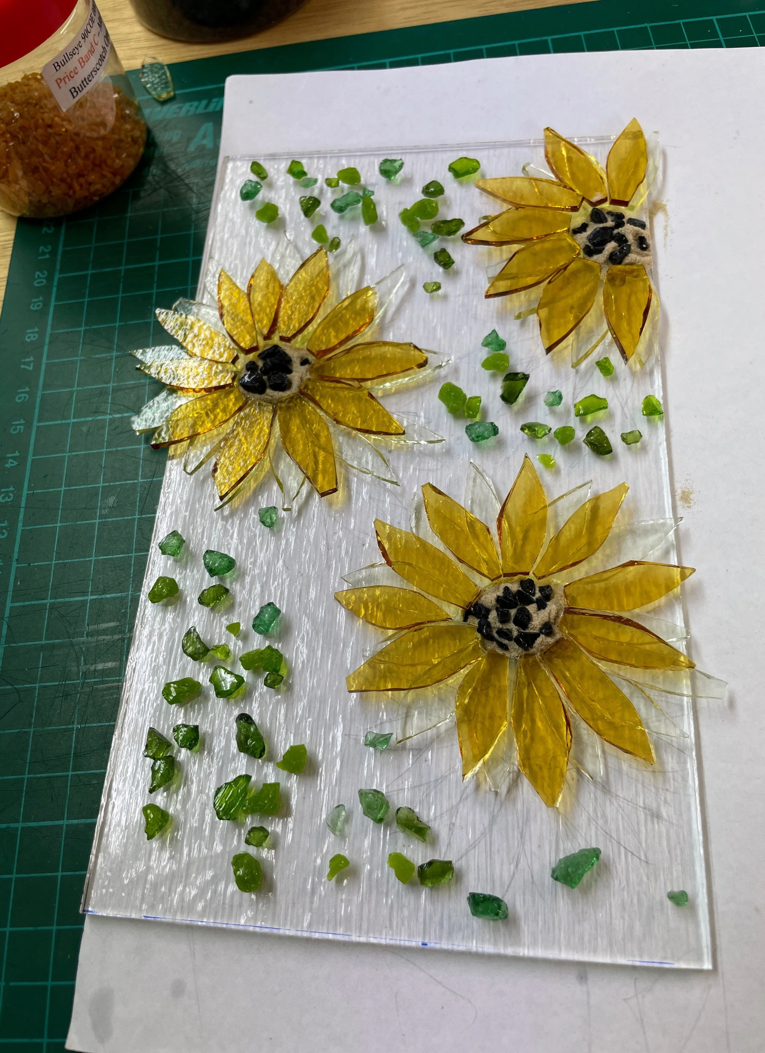 Glass mosaic art featuring sunflowers and green leaves on a glass panel, with yellow and green glass pieces used for designs, set on a green cutting mat.