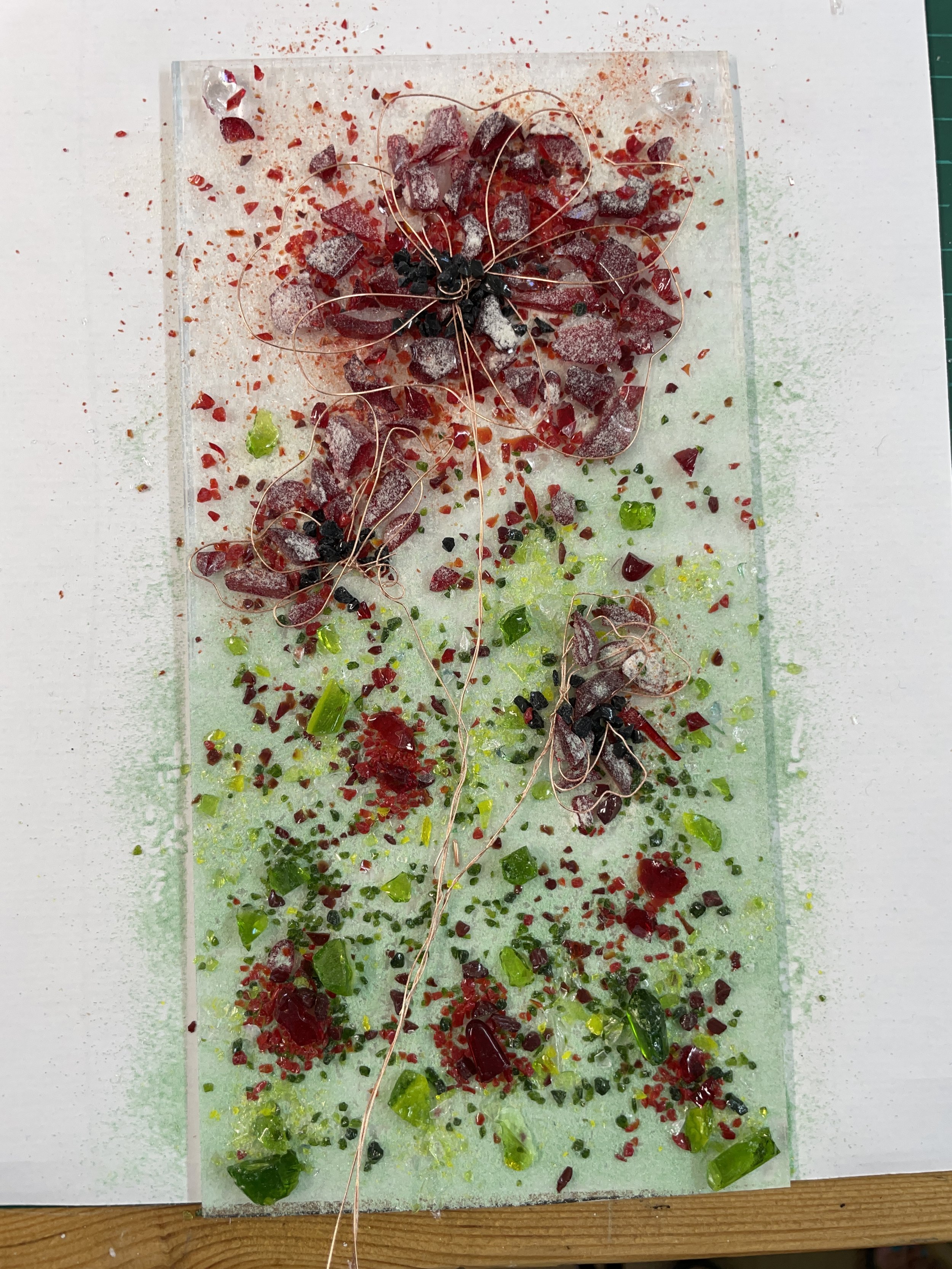 Glass art piece featuring red and green flower designs with wire stems and scattered glass fragments on a light background.